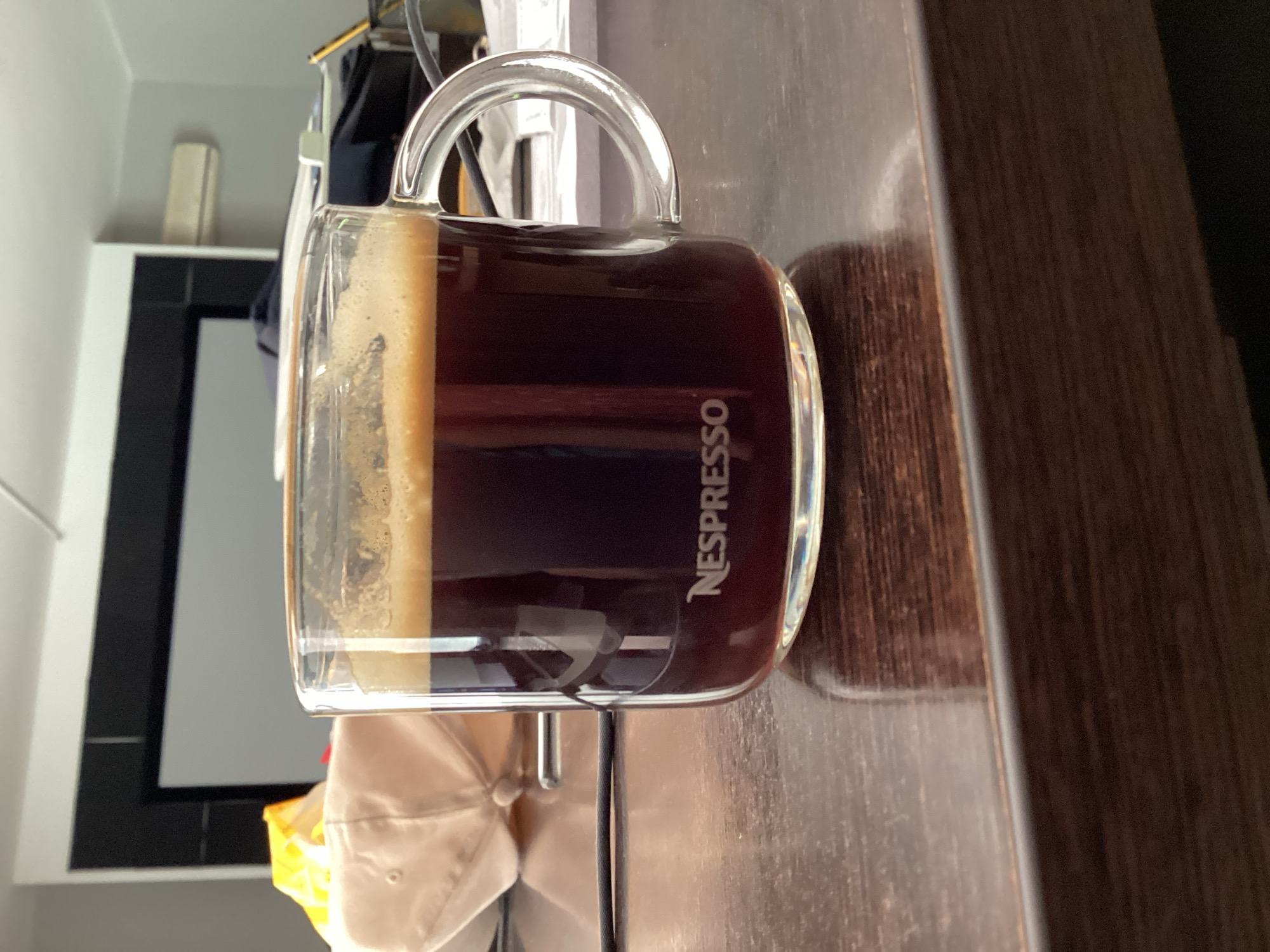 A close-up, eye-level shot shows a clear glass mug filled with dark coffee and a layer of crema. The mug is emblazoned with the white "NESPRESSO" logo. It rests on a dark, wooden surface, likely a desk or table. In the background, a white baseball cap and a yellow object are partially visible, suggesting a casual setting. The background also features a large, dark-framed screen or projector, and a white air conditioning unit mounted on a light gray wall, indicating an indoor environment. This scene could be located anywhere, but if specifically requested to be in Blanes, Spain, the location would be an indoor space within the city.