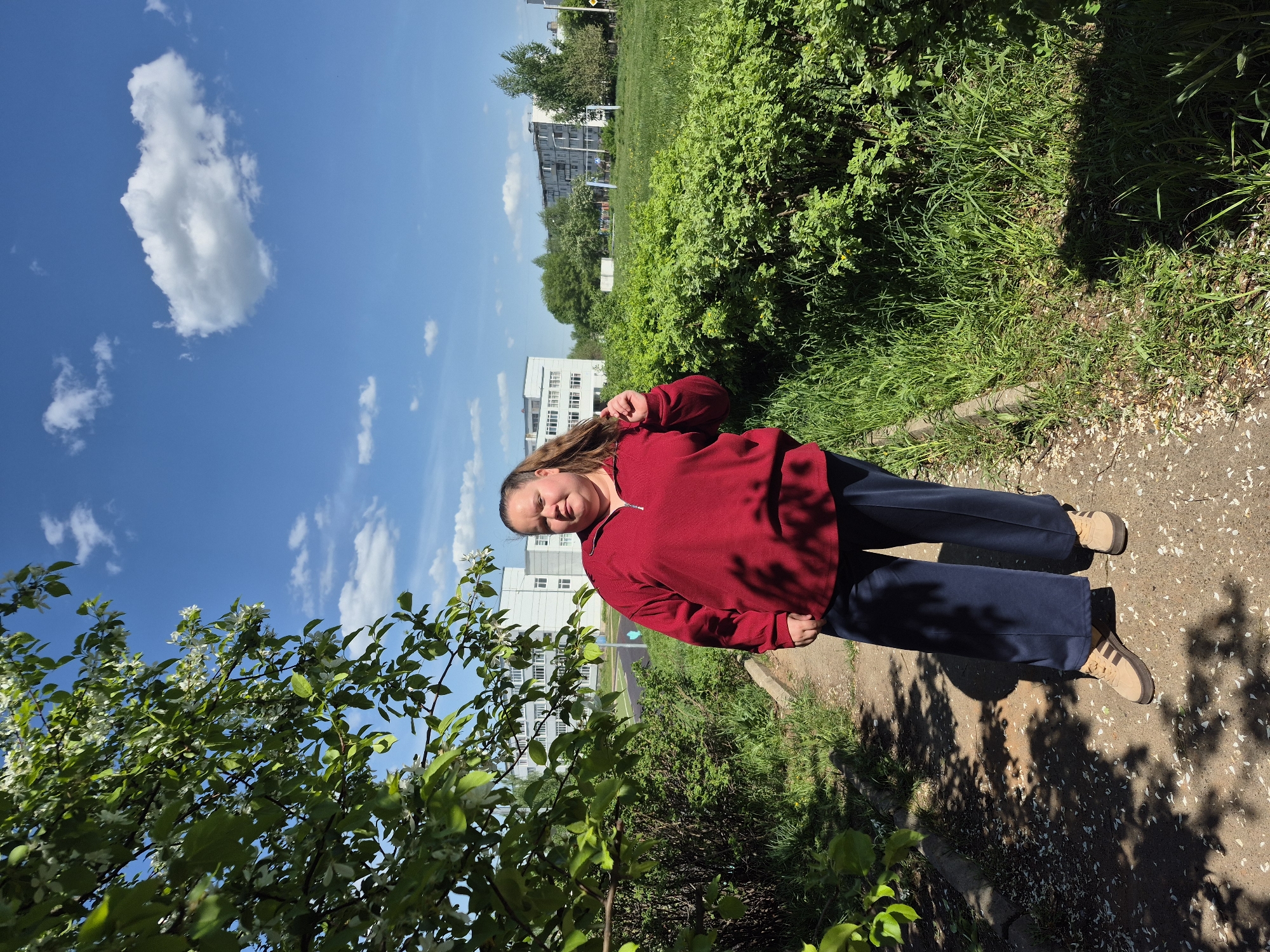 A woman stands on a sidewalk in Chaikovsky, Russia. She has brown hair, a red sweater, navy pants, and tan shoes. A building stands in the background to the left, with trees and bushes. The sky is blue with clouds.