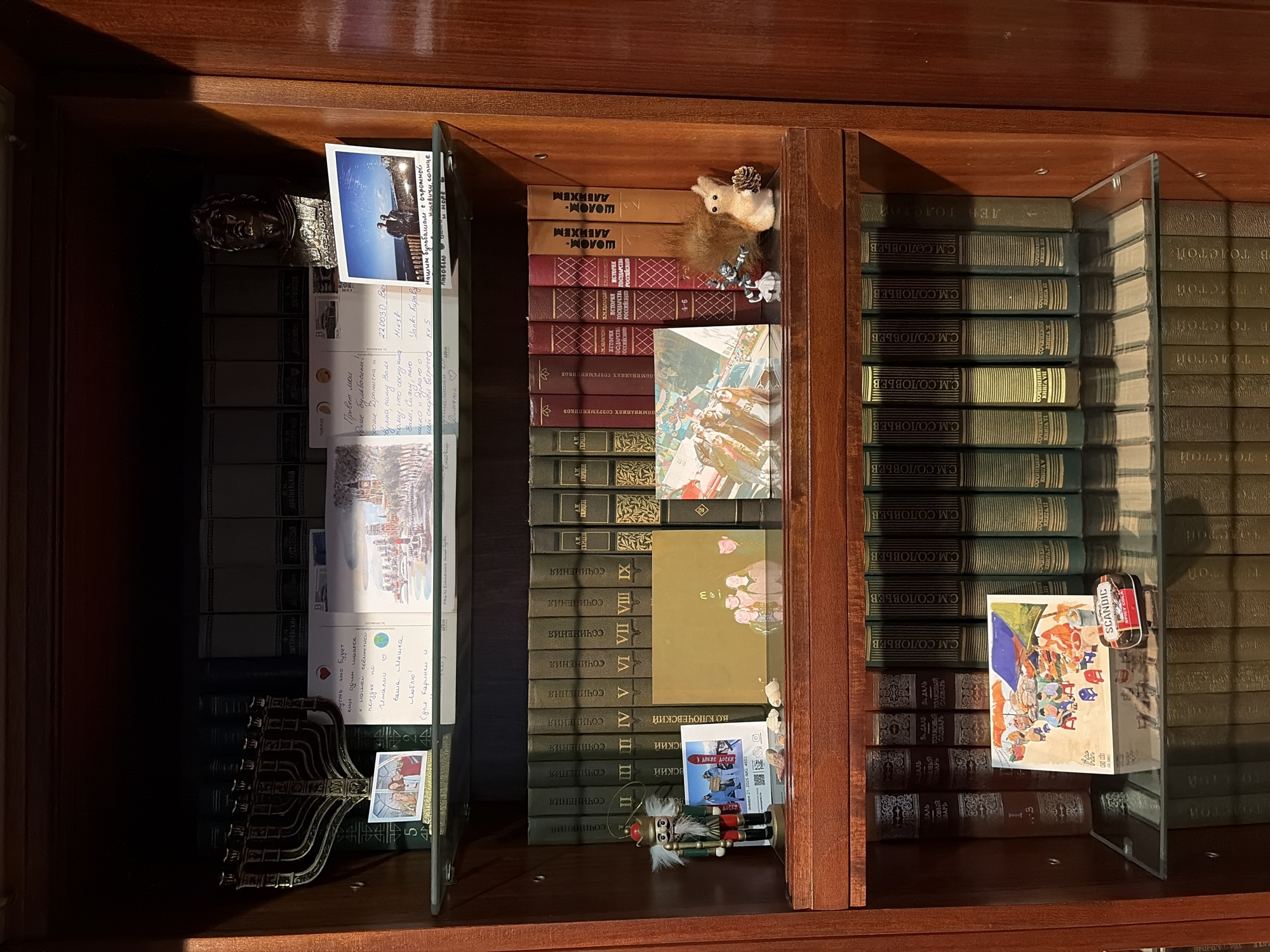 A photograph of a wooden bookshelf containing books and other objects. The bookshelf is divided into three sections, each with books stacked on top of each other. The books are mostly old and have titles written in Russian. In the left section of the bookshelf, there are several postcards and letters leaning against the books. There is also a decorative object that looks like a menorah. In the center section of the bookshelf, there is a stuffed animal squirrel and a small nutcracker figurine. There is also a postcard with a picture of people sitting at tables under umbrellas. In the right section of the bookshelf, there is a postcard with a picture of a group of people sitting around a table. There is also a small tin of Swedish candy. The city and state are not specified.