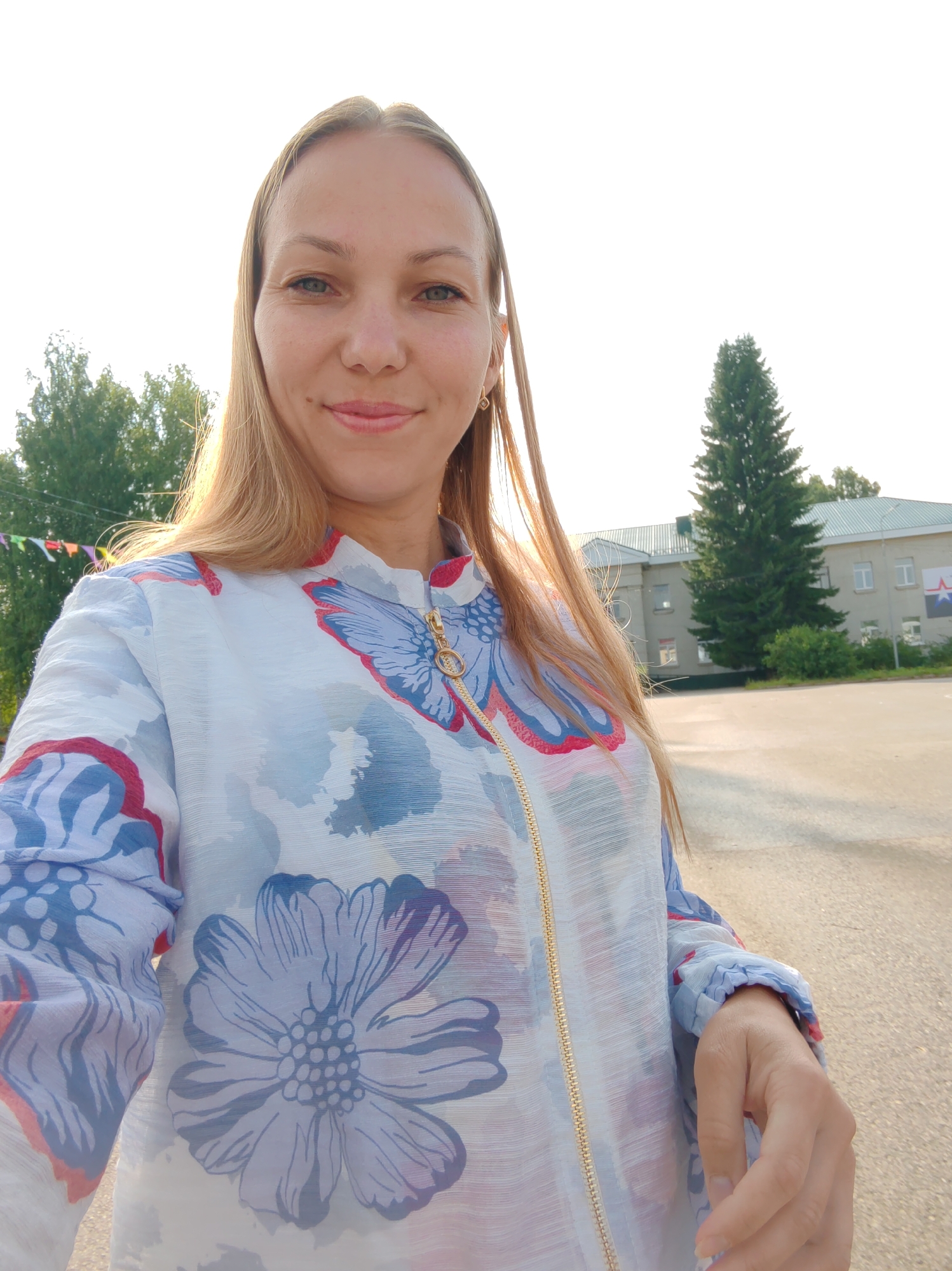 In Vizinga, Russia, a woman with blonde hair, wearing a light-colored shirt with a floral pattern, takes a selfie outdoors. The background includes trees, a building, and a clear sky. The woman is smiling and looking at the camera. The lighting suggests it is daytime, possibly late afternoon due to the warm glow. The environment appears to be a public space, perhaps a park or a street near a building.