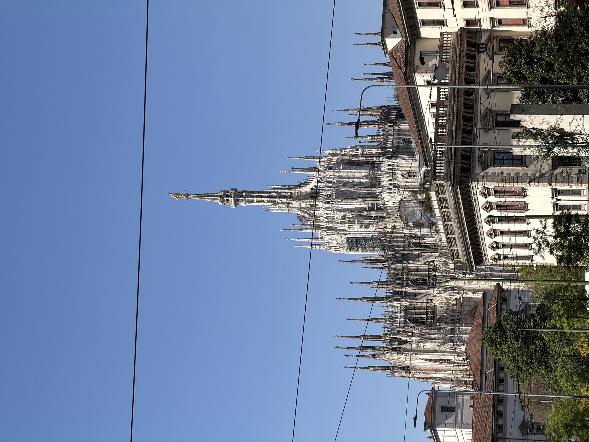 The image depicts the Milan Cathedral, a prominent landmark in Milan, Italy. The cathedral's intricate facade, characterized by numerous spires and statues, dominates the scene. The architecture is Gothic in style, suggesting the cathedral's historical and cultural significance. A clear blue sky forms the backdrop, enhancing the cathedral's silhouette. Power lines are visible in the foreground, creating a contrast between modern infrastructure and historical architecture. Adjacent buildings are seen alongside trees.