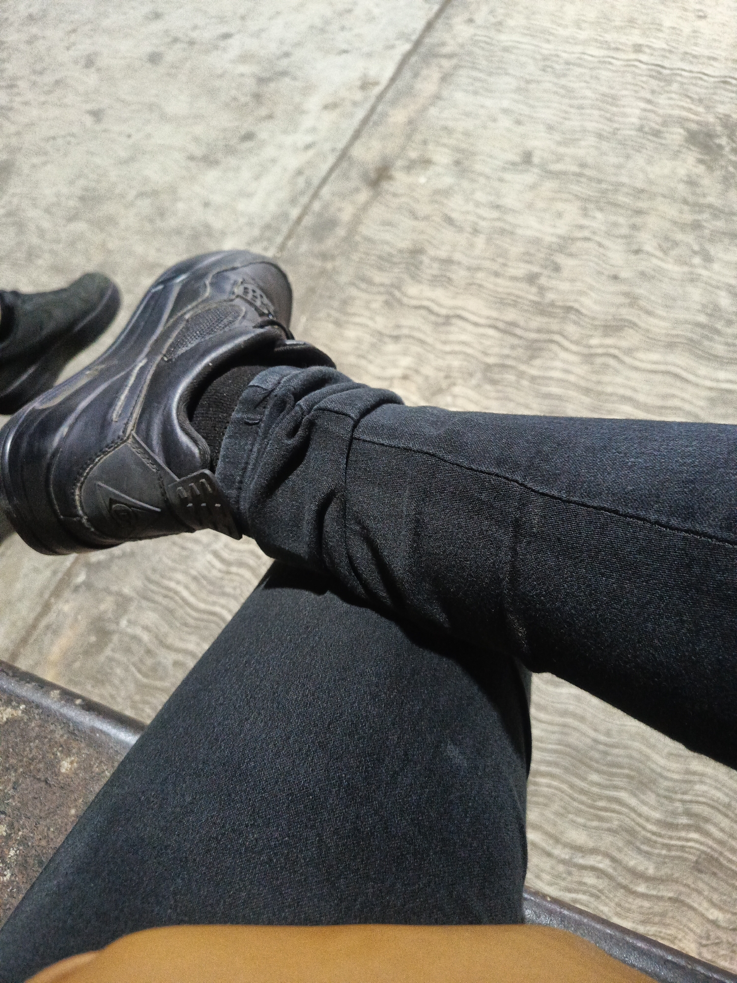 A high-angle, close-up shot shows a person's crossed legs, clad in dark denim jeans, with black sneakers on their feet. The legs are resting on what appears to be a bench or ledge. The surface beneath the feet is a light-colored concrete with a textured, linear pattern. The scene provides no discernible cues to suggest a specific location within Juan Griego, Venezuela.