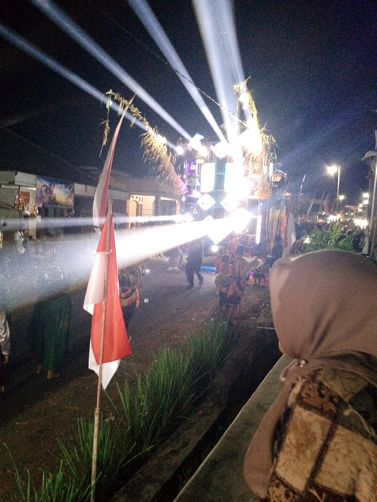 The image captures an outdoor nighttime scene in Babadan, Indonesia, likely a festive gathering. Bright white spotlights cut through the darkness, illuminating a stage or performance area in the background, where several figures are visible. A prominent Indonesian flag, red and white, stands in the foreground, partially obscuring the view of a crowd of people in the middle ground. Some individuals appear to be dancing or moving. In the immediate foreground, a person wearing a head covering and a patterned garment is partially visible, facing away from the camera and looking towards the event. The ground is dirt or gravel, with some green vegetation, possibly grass, scattered throughout. Buildings and streetlights are faintly visible in the distance.