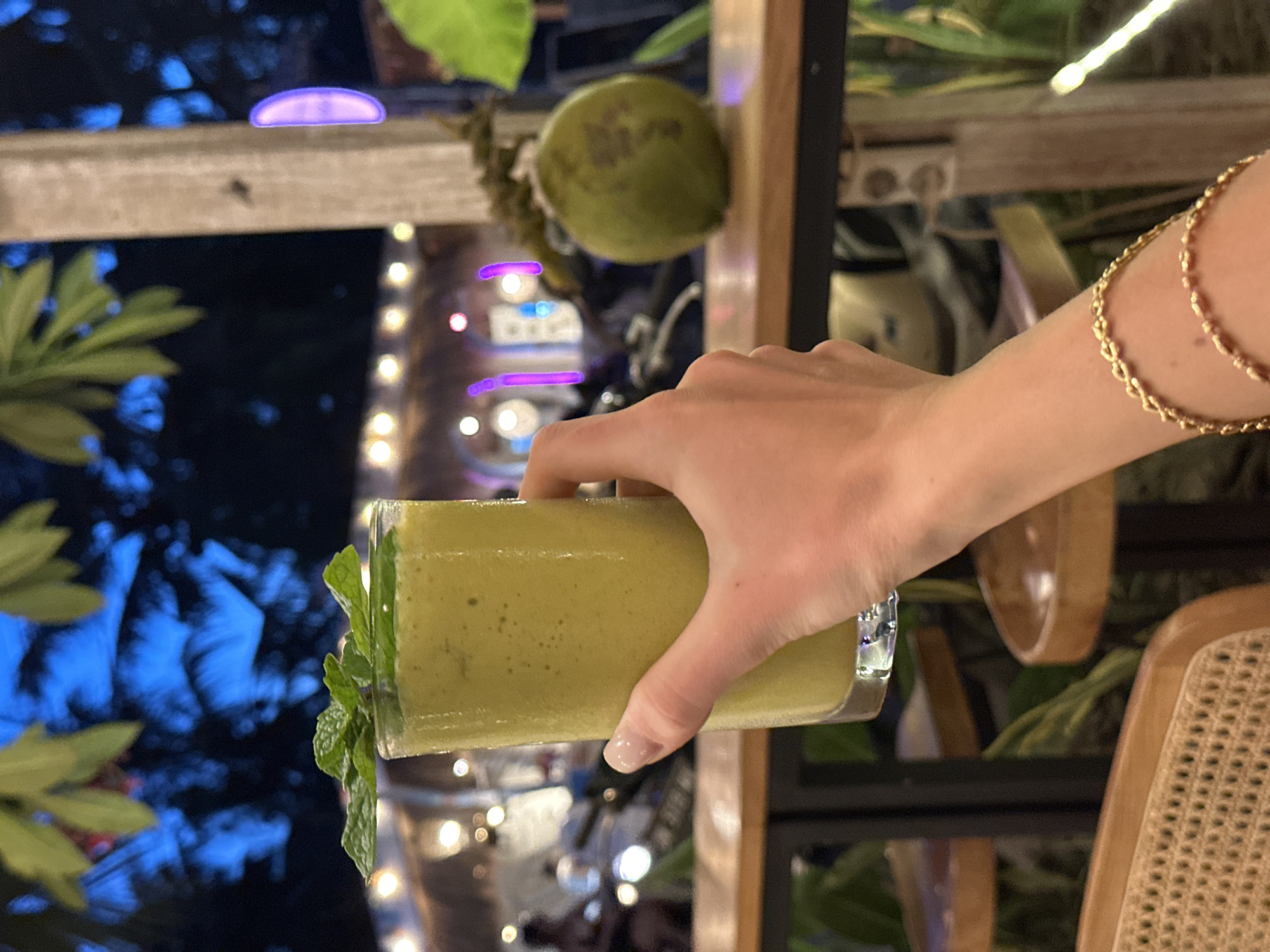 A person holds a glass of green smoothie in their right hand in a restaurant in Nusa Penida, Indonesia. The hand is adorned with gold bracelets. The drink is garnished with mint leaves. A young coconut hangs behind the glass. Outside the window, blue and white decorative lights are visible among trees. A rattan chair is nearby. The restaurant is decorated with plants.