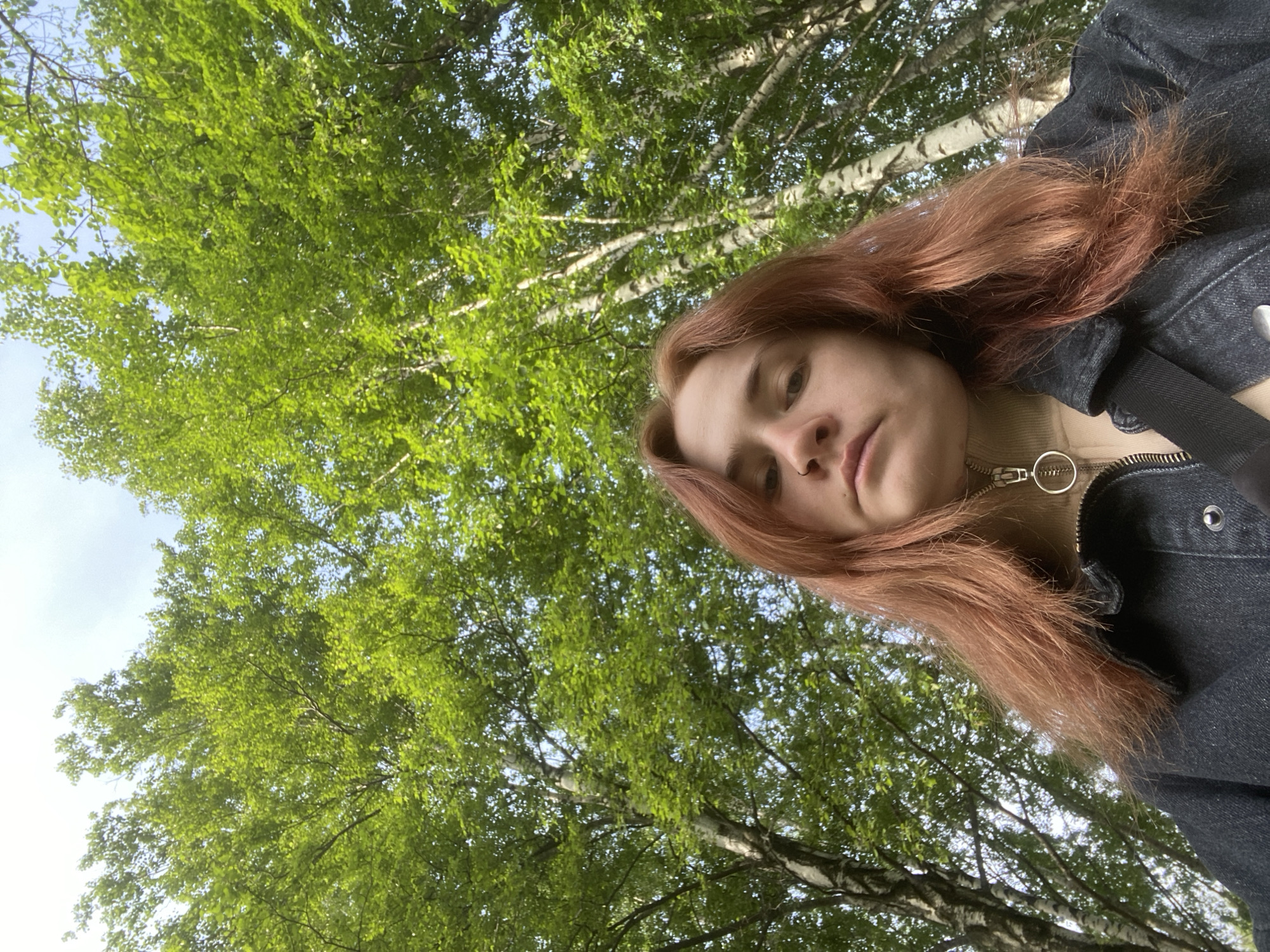 A woman with reddish-brown hair is visible in the foreground, wearing a dark denim jacket over a lighter-colored turtleneck sweater with a ring zipper pull. The background shows a dense canopy of green leaves and white-barked trees, suggesting a forest or park setting. Based on the prompt, the location is presumed to be within the city of Chaikovsky, Russia.