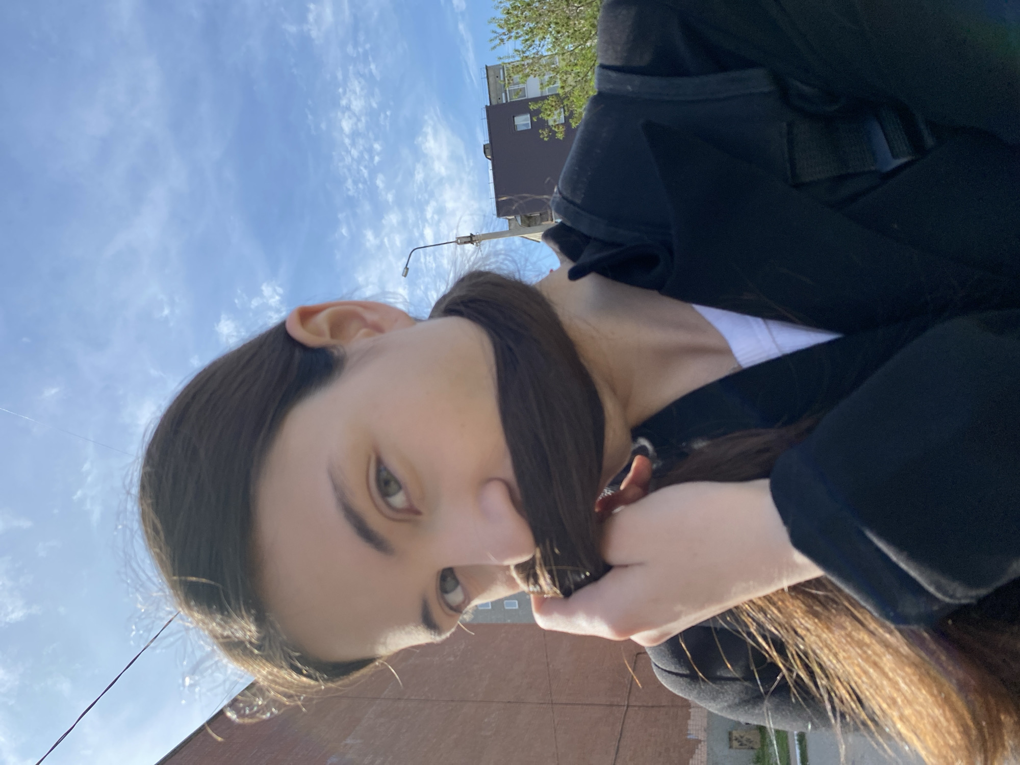 A low-angle selfie taken in an urban setting, likely Votkinsk, Russia, based on architectural styles. The primary subject is a young woman with long brown hair, part of which she holds in front of her face, partially obscuring it. She has fair skin and green eyes. She is wearing a white top underneath a black jacket. A portion of her backpack is visible on her right shoulder. The background includes a light blue sky with scattered clouds, a section of a brick building, a streetlight, a brown building, and green trees. The camera angle suggests the person is looking down slightly.