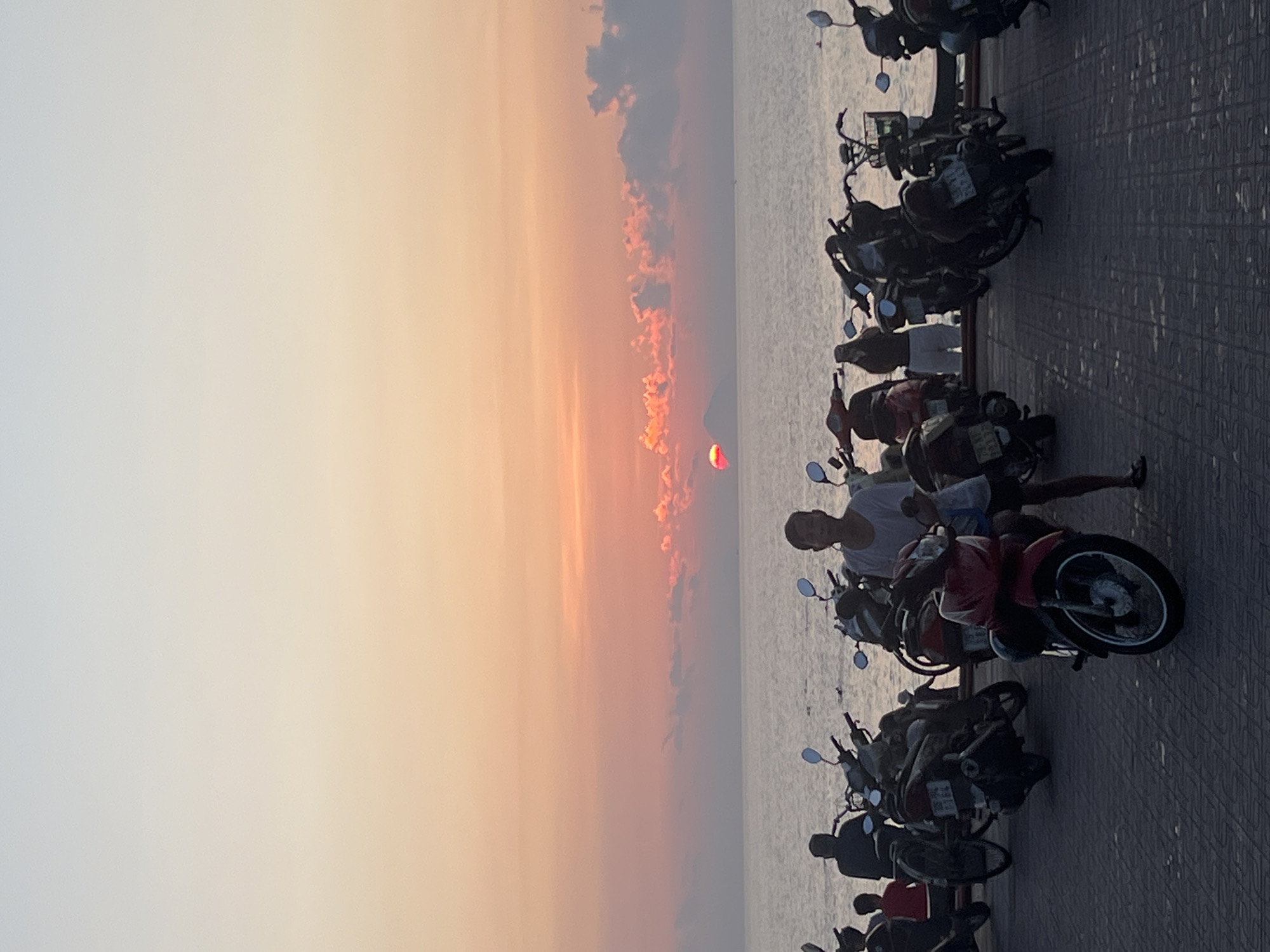 The image captures a scenic sunset over a calm body of water, likely the sea, in Nha Trang, Vietnam. The time of day is late afternoon or early evening, indicated by the vibrant orange, red, and yellow hues dominating the horizon as the sun sets. The weather appears clear with scattered, wispy clouds reflecting the sun's colors.
In the foreground, a paved promenade, possibly a boardwalk or seawall, runs parallel to the water. A long line of parked motorbikes and scooters occupies the right side of the image, facing the sea. These vehicles are primarily dark and silhouetted against the brighter background, with some details discernible, such as handlebars, mirrors, and various parts of their frames.
Several people are visible among the motorbikes. One individual in a light-colored top is prominently seated on a red scooter, looking towards the left, with their legs down. Another person in a light shirt is standing near a scooter behind them, partially obscured. Further down the line, other figures are present, mostly as indistinct silhouettes, suggesting people gathered to observe the sunset.
The scene depicts a leisurely moment, characteristic of a popular coastal area where individuals congregate to enjoy the evening view. The sun, a distinct red-orange disc, is partially visible above the horizon, casting a warm glow across the sky and reflecting subtly on the water. No readable text is visible on the motorbikes or elsewhere in the image.