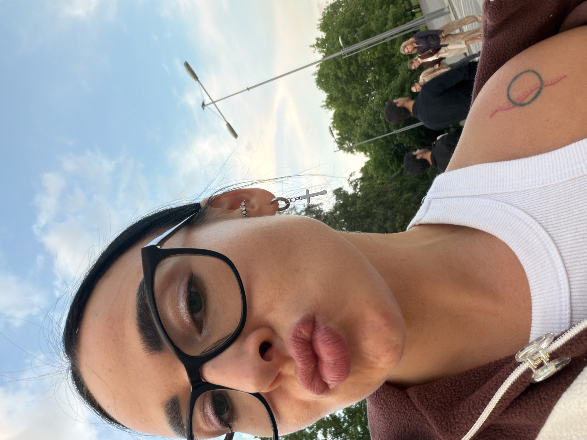 The image is a close-up, low-angle selfie of a young woman, looking upwards, capturing her face and shoulder against the sky. The woman has dark hair and wears black-rimmed glasses. She makes a playful, pouty expression with her lips. Her left ear features multiple piercings, including a silver cross charm dangling from a hoop earring and a small, jeweled ear cuff. She wears a white ribbed tank top underneath a brown, possibly fleece, zippered jacket with a translucent, shell-like zipper pull. On her right upper arm, a tattoo depicts a blue circle with a red line extending from it.
In the blurred background, several other individuals are visible, out of focus, suggesting an outdoor public space. Green trees and streetlights extend into the frame, set against a soft blue sky with scattered white clouds. The diffused lighting suggests late afternoon or early evening. The scene is likely an urban or park-like environment in Saint Petersburg, Russia, capturing a casual moment. No visible text is present.