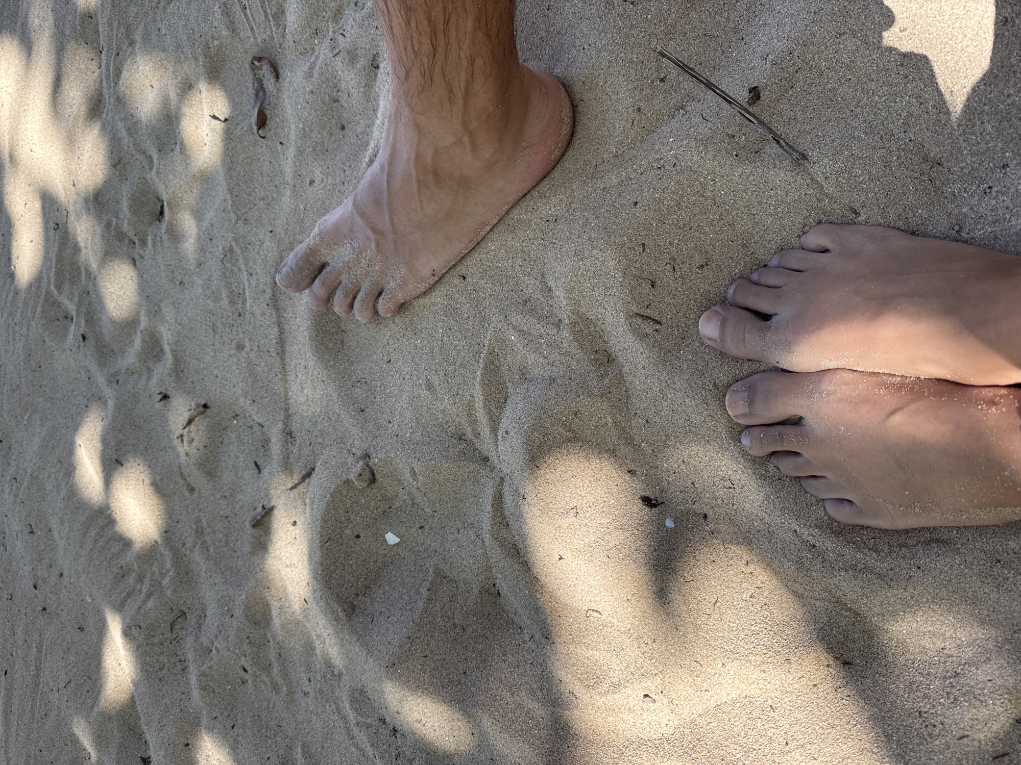 This image depicts a sandy beach in Kuta Selatan, Indonesia. Two pairs of bare feet are partially visible on the sand. One pair is lighter in skin tone and positioned close together; the other is darker, with the lower leg visible. Scattered twigs are on the sand. Patches of sunlight dappled through foliage cast shadows across the scene.