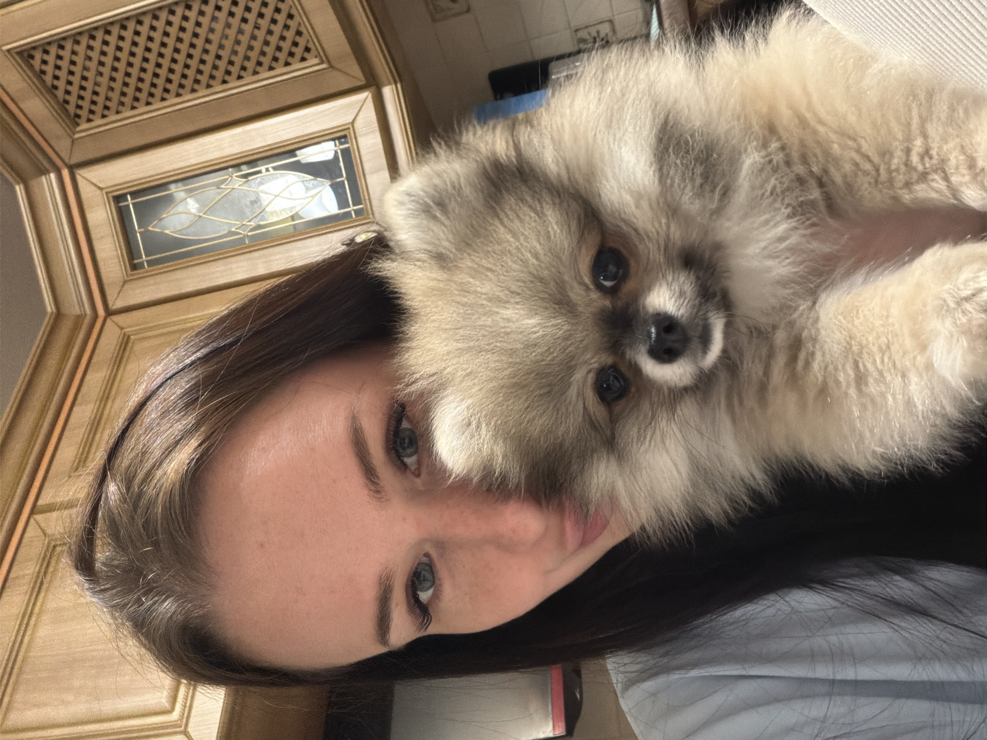 This media file presents a close-up portrait featuring a young woman and a small dog indoors. The image is captured with a vertical orientation, showing the subjects from an overhead perspective, with the background appearing to the left.
The primary subjects are a woman with long, dark brown hair and clear blue eyes, and a fluffy, light brown and grey Pomeranian dog. The woman's face is visible on the left side of the frame, looking directly at the camera with a neutral expression. The dog is positioned beside or on her, with its head resting near her face, also looking straight into the lens. It has dark, expressive eyes and a small black nose, characteristic of its breed.
The background details suggest an indoor home environment. To the left, there is elaborate wooden cabinetry, possibly in a kitchen or living room. The top section of the cabinetry features a decorative latticework panel. Below this, another cabinet door incorporates a rectangular glass insert, behind which a light fixture with a leaf-like design is visible. The wood has a warm, natural tone. The lighting appears soft and natural, suggesting it is daytime. No text is visible in the image.