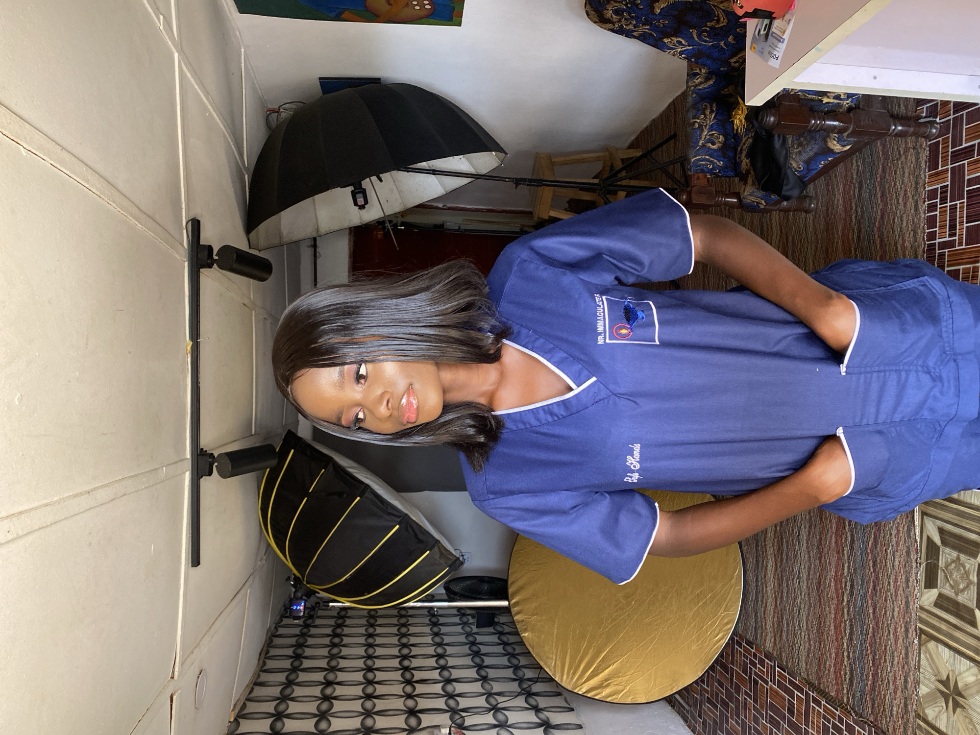 The image captures a young woman posing indoors, likely in a photography studio in Vom, Nigeria. She stands with her hands in the pockets of her blue scrub-like uniform, looking towards the camera with a calm, slightly smiling expression. Her dark, shoulder-length hair is neatly styled. The uniform features white piping along the V-neck collar and cuffs. On the left chest, "NRI. IMMACULATE" is embroidered, accompanied by a logo displaying a blue silhouette within a red circle, outlined in white. On the right side of the uniform, the words "Bulu Hands" are embroidered in a script font.
The background shows various pieces of photography equipment, including a large octagonal softbox on a stand to her left, a smaller rectangular softbox to her right, and a gold circular reflector resting on the floor. The setting appears to be an interior space with mixed wall and floor coverings; a light-colored panel wall is visible on the left, while the floor exhibits a dark geometric tiled pattern, a striped rug, and brown/beige tiles. The artificial lighting suggests the time of day is not a factor.