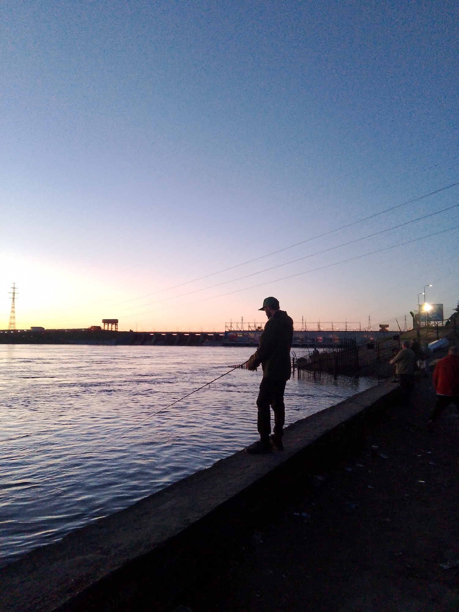 A man is fishing at sunset in Chaikovsky, Russia. He stands on a concrete barrier along the bank of a wide river, holding a fishing rod. Other figures stand behind him further along the embankment. In the background, across the river, is a large industrial structure with a tower on the left. Power lines are visible in the upper right. The sky is a gradient of blue fading to orange at the horizon. The water reflects the colors of the sky. The overall impression is one of peaceful evening activity.