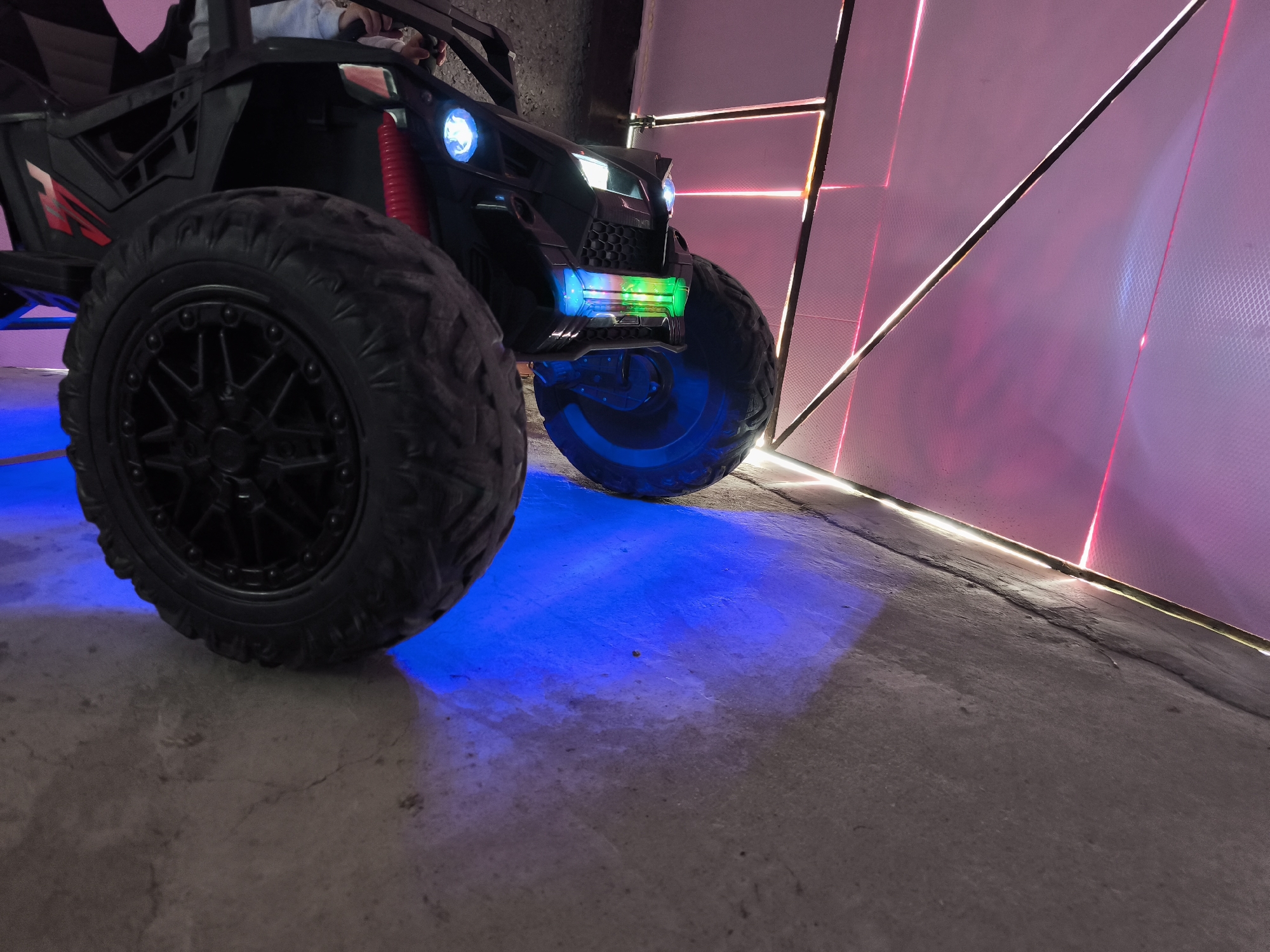 A low-angle shot captures a black children's electric ride-on car positioned indoors against a backdrop of a pink geometric wall. The car features large, knobby black tires and illuminated headlights in a bright white-blue hue. The front of the vehicle is accented with a multi-colored light strip consisting of blue, green, and yellow tones. The floor, made of concrete, is illuminated with a blue light emanating from the car. The background wall is a grid of pink-colored panels with black framing.