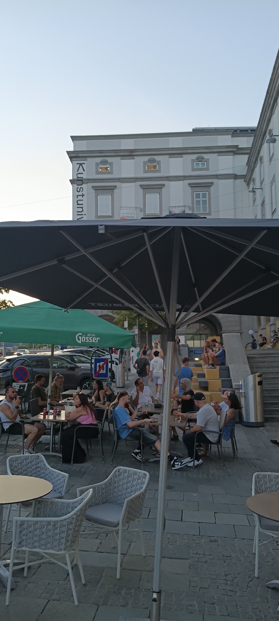 The image depicts an outdoor seating area of a cafe or restaurant in Linz, Austria, likely near a public building. In the background, a large, light-colored building with classical architectural elements is visible, featuring rows of windows and columns. A prominent sign on the building reads "Kunstuni," indicating it is likely the University of Art and Design Linz.
In the foreground, a large black umbrella shades tables and chairs occupied by patrons. Several people are seated at these tables, engaged in conversation or enjoying drinks and food. The patrons are diverse in age and gender, dressed in casual attire. Some individuals are smoking, and drinks such as beer are visible on the tables.
To the left, a green umbrella with the "Gösser" beer logo is also visible, providing shade to another section of the seating area. Cars are parked behind this area, and signage, including a no-parking symbol, is present.
To the right, a set of tiered outdoor steps leads up to an entrance, with more people seated on the steps. The overall atmosphere suggests a relaxed, social setting, likely during the late afternoon or early evening, given the lighting conditions. The ground is paved with cobblestones.