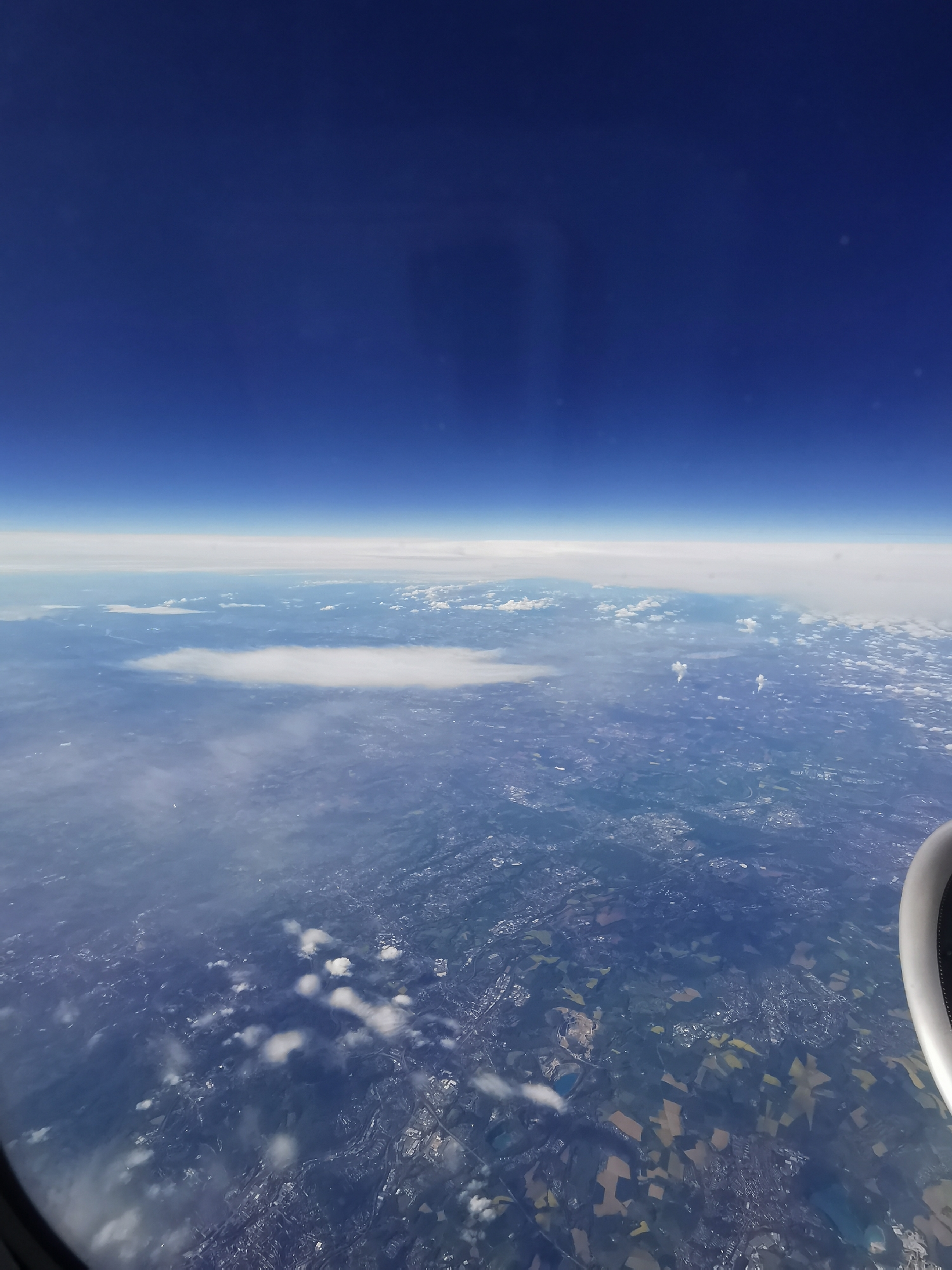 The image shows a view from an airplane window. The sky is a clear, deep blue. Below the horizon is a layer of white clouds, and further down, the earth is visible, showing a mix of fields, possibly agricultural lands, and what could be urban or developed areas. In the bottom right corner, a part of the airplane's engine is visible. It's impossible to determine if the location is in Varna, Bulgaria without additional information.