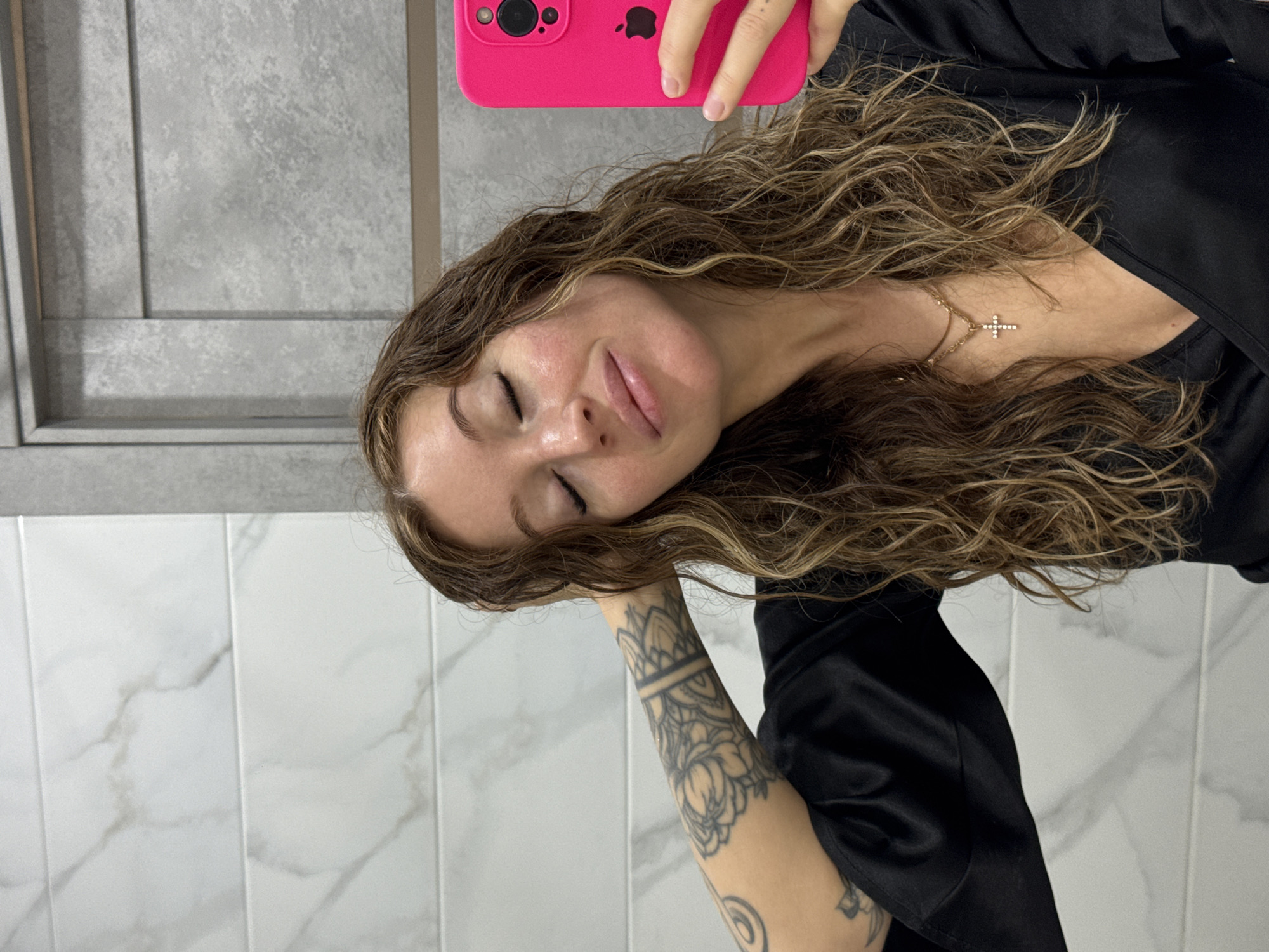 The image is a selfie of a young woman, taken indoors in Chaikovsky, Russia. She is shown with her eyes closed, resting her head on her left arm, which is extensively tattooed with intricate, dark patterns, possibly floral or mandala-inspired designs. She has long, wavy, brown hair with lighter highlights, cascading around her shoulders. She wears a black garment, resembling a satin robe or top, and a gold-colored chain necklace with a small, sparkly cross pendant.
She holds a bright pink smartphone, visibly an iPhone due to the distinct camera module and the Apple logo on its back, to capture the image. Her expression, with eyes closed and a slight smile, conveys a sense of calm and relaxation.
The background features two different types of wall surfacing. The upper part consists of a light gray, textured wall with subtle horizontal and vertical panel lines. The lower part of the background is composed of white, rectangular tiles with light gray, marble-like veining, suggesting a modern interior space, possibly a bathroom or a dressing room. The lighting appears to be artificial, typical of an indoor setting. There is no visible text apart from the Apple logo on the phone.