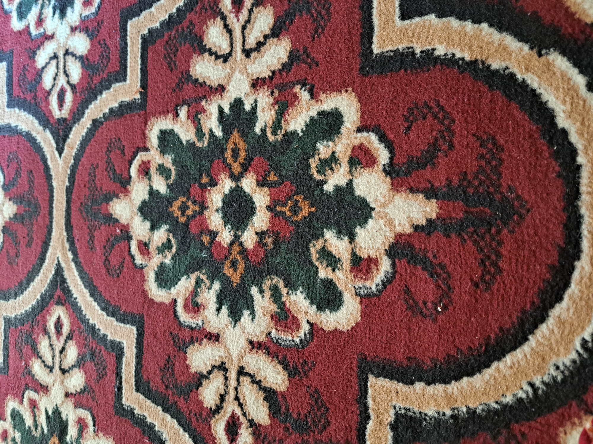 The image displays a close-up view of a Persian-style carpet. The carpet features intricate floral patterns in shades of deep red, cream, black, and green, with small accents of orange and yellow. The design is symmetrical, with a central motif that is surrounded by a larger, scrolling border. The texture of the carpet appears plush and soft. The lighting suggests an indoor environment, likely a room within a building. This carpet is situated in the city of Qarnon, Uzbekistan, specifically within a room. There are no people or actions depicted in the image.