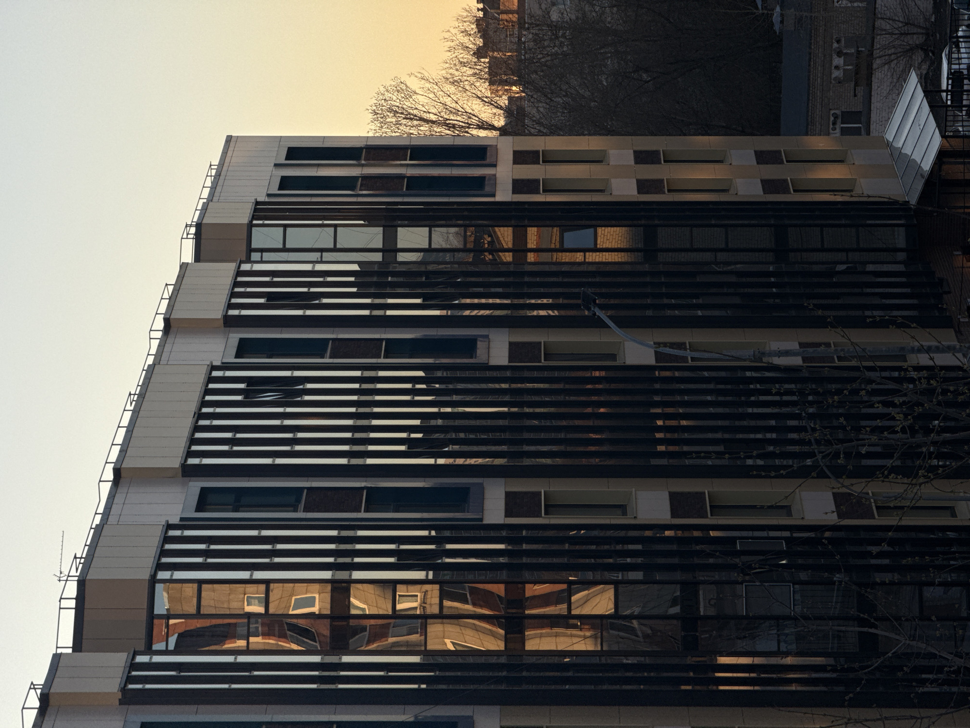 The image shows an exterior view of a multi-story building with a modern architectural design. The building is characterized by horizontal rows of windows separated by dark, linear elements, possibly metal or composite material. The windows reflect images of surrounding buildings, suggesting a dense urban environment. The building's exterior consists of light-colored panels, possibly stone or concrete. The image is shot at an oblique angle, and the sky is visible at the top, with a light, hazy quality. Bare tree branches are partially visible in the foreground and top of the image. Based on the prompt's information, the location is likely in Perm, Russia.