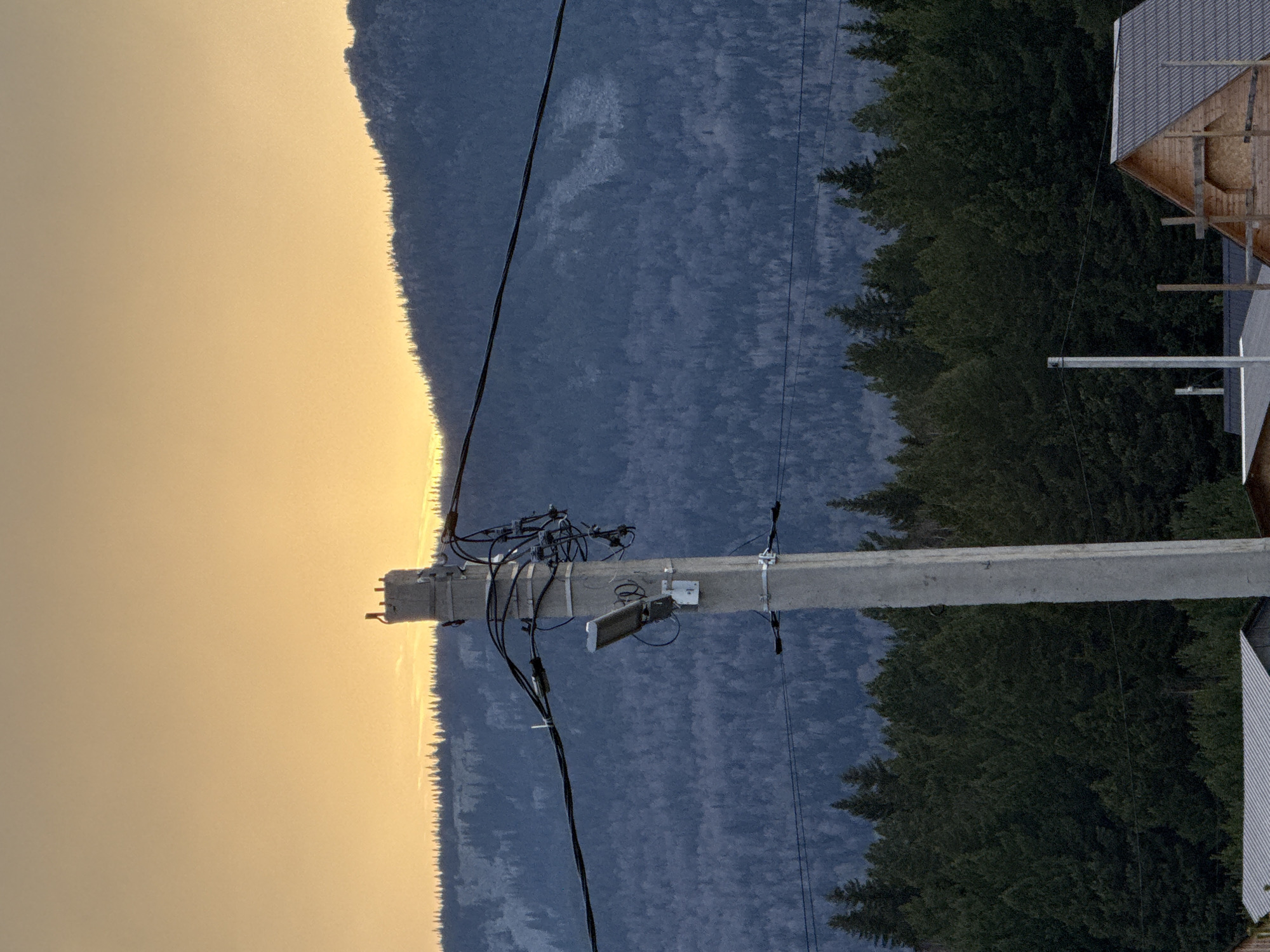 The image shows a utility pole situated in what appears to be the rural setting of Tyulyuk, Russia. The pole is concrete and supports electrical wires and some equipment including what appears to be a streetlight fixture. In the background is a mountain range shrouded in a bluish-grey haze, with a band of trees separating the mountains from the sky. The sky is a pale yellow, suggesting the time of day is either sunrise or sunset. A partially-constructed wooden building, likely a residential structure, is visible on the right side of the frame.