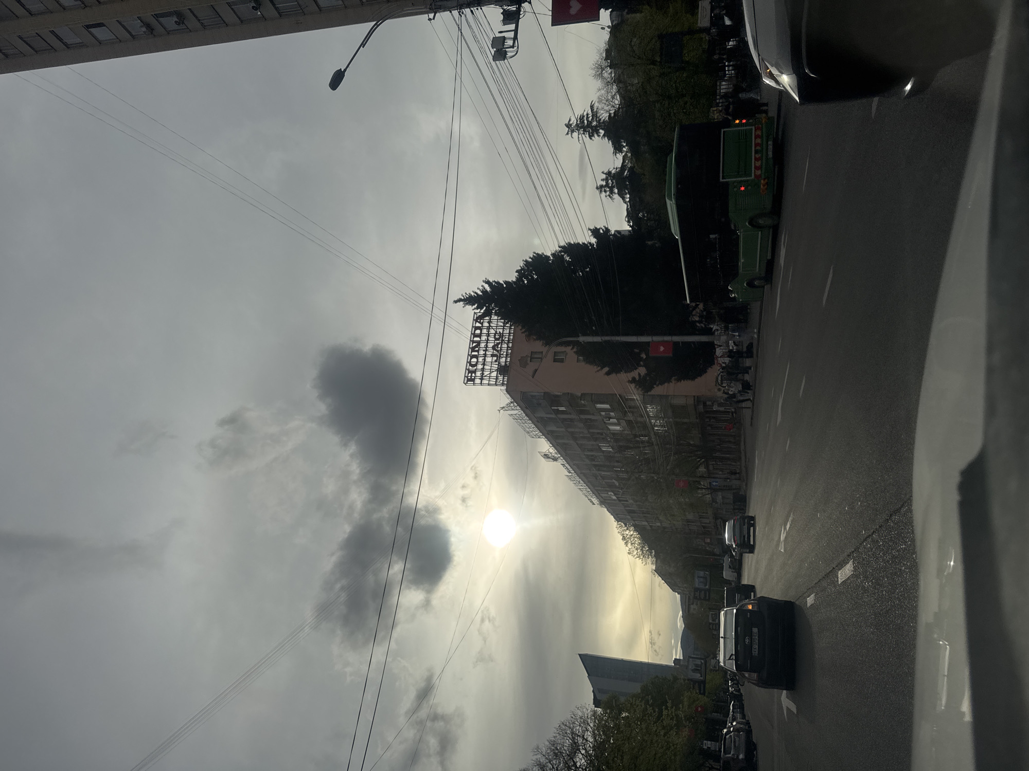 This media file captures an urban street scene in Tbilisi, Georgia, under an overcast sky with the sun piercing through the clouds. The image is taken from within a vehicle, looking towards the street and buildings.
The sky is largely cloudy but brightened by the visible sun, which casts a strong glare. Numerous utility wires crisscross the frame against the cloudy backdrop. On the right, an asphalt road features white lane markings and directional arrows. Several vehicles are present: a dark sedan with a Georgian license plate (partially visible as "XYL-001" or similar), a green vehicle resembling a bus or food truck, and the front of a light-colored vehicle in the immediate foreground.
Multi-story buildings line the street. One prominent building features a large, metal framework sign on its roof, clearly displaying the text "HONDA". Another building has what appears to be scaffolding or an architectural detail on its upper structure. Trees, some with bare branches and others with foliage, are scattered along the sidewalks and between buildings. Streetlights and traffic signal poles are also visible, some adorned with small red signs featuring a white heart symbol.
The scene depicts typical daytime urban activity, with vehicles navigating or parked on the street. The light suggests it is late morning or afternoon.