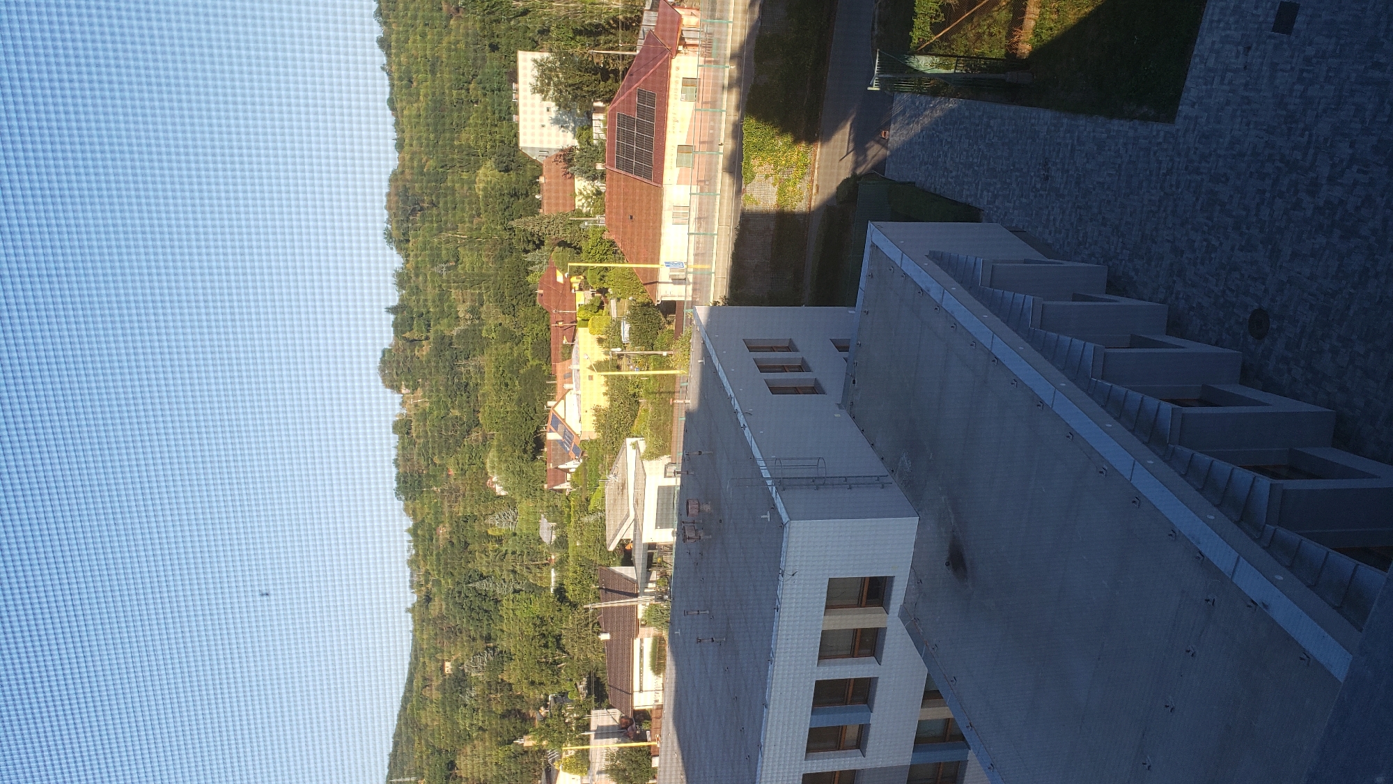 The image is a high-angle shot of a suburban area with buildings and a wooded hill in the background. In the foreground, there are two modern, flat-roofed buildings. The building on the left has a light gray facade with horizontal rows of windows. The building on the right, partially visible, has a gray roof and a walkway leading to it. In the middle ground, a street runs horizontally, bordered by a fence. Across the street, there are several residential houses with reddish-brown roofs. One house has solar panels on its roof. The background features a densely wooded hill with scattered houses visible amongst the trees. The sky is clear and blue, with a subtle grid pattern, suggesting the image was taken through a screen or window. There are no people visible in the image. The scene appears to be a typical residential and institutional area in a hilly landscape.