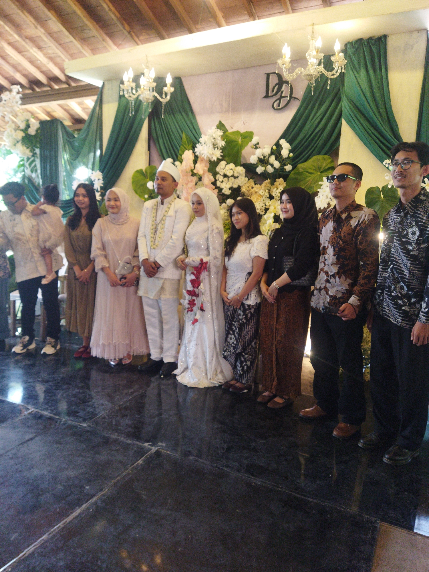 The image depicts a formal gathering, possibly a wedding reception, set against a backdrop of floral arrangements and draped green fabric. Several individuals are lined up, appearing to pose for a photograph.
In the foreground, the polished dark floor reflects the overhead lighting. The individuals are dressed in a mix of traditional and modern attire.
From left to right:
The first person is a male wearing a white shirt and black pants, holding a child. The child is wearing a light-colored outfit.
The second person is a female in a long, olive-green dress.
The third person is a female in a pale pink, floor-length dress and a light pink headscarf. She is holding a small silver purse.
The fourth person is a male in a white traditional suit, complete with a white head covering and a beaded necklace.
The fifth person is a female, the bride, wearing a white, intricately beaded wedding dress and a white veil.
The sixth person is a female wearing a black hijab and a black top over a patterned skirt and a brown batik sarong.
The seventh person is a male wearing a patterned shirt and dark pants, with sunglasses on.
The eighth person is a male wearing a dark patterned shirt and black pants.
The ninth person is a male wearing a dark patterned shirt and black pants.
The background features a wall with a stylized "DD" monogram. Green drapes are strategically placed, framing the scene. Chandeliers with multiple light bulbs hang from the ceiling, which appears to be made of wood. The overall ambiance suggests an indoor event, possibly in a venue in Semarang, Indonesia, given the cultural attire and decorative elements commonly seen at Indonesian celebrations.