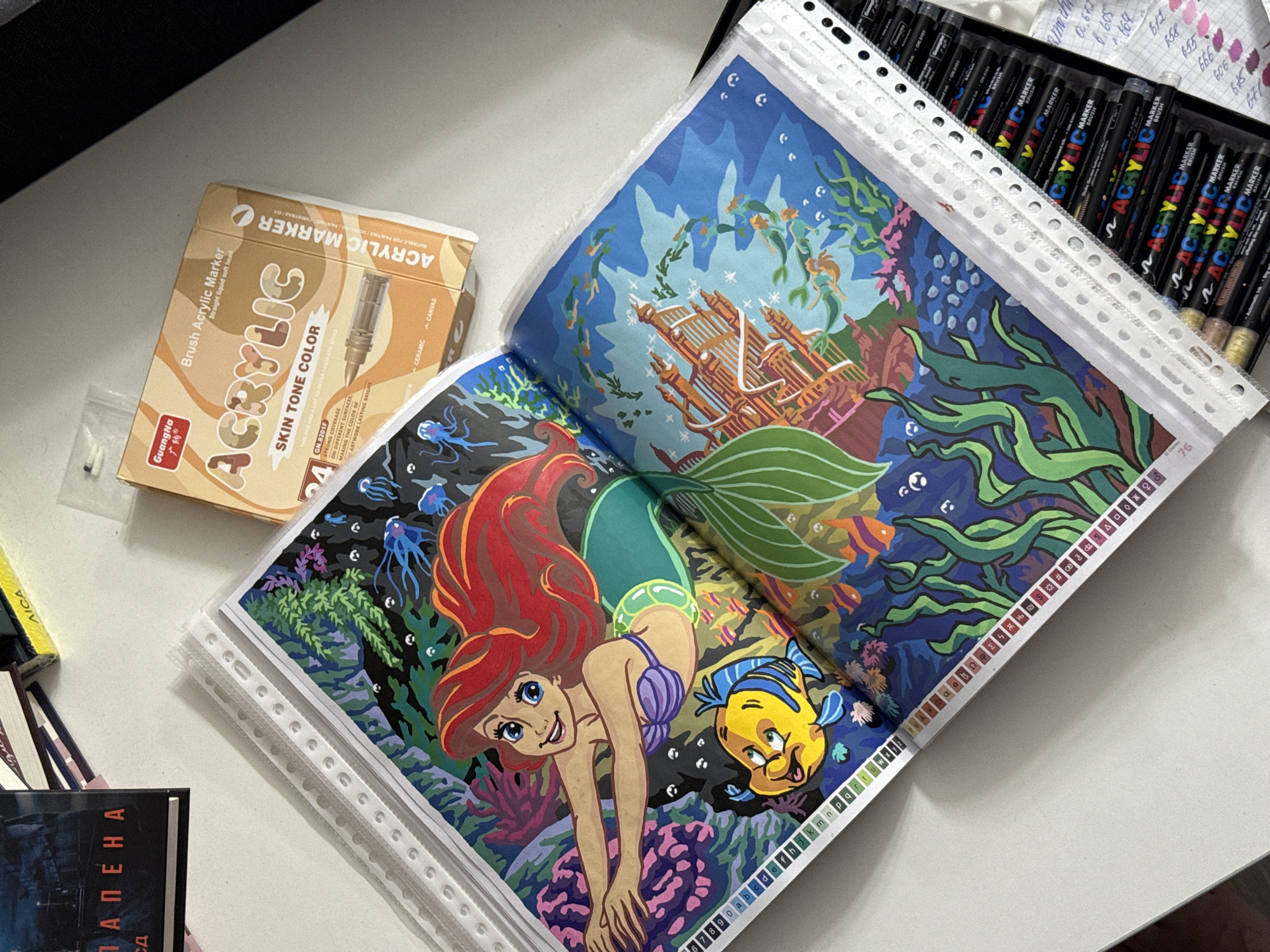 The image shows a top-down view of a table with art supplies. In the foreground, an open coloring book features a colorful illustration of Ariel and Flounder from Disney's The Little Mermaid, set in an underwater scene with a castle. The coloring book pages are protected in plastic sleeves. To the right of the coloring book, a box of "GuangNa Acrylic Marker" pens, specifically labeled "SKIN TONE COLOR," is visible. Below this box, a collection of various "ACRYLIC MARKER BRUSH" pens are neatly arranged in a case, with some of their caps removed. In the upper left corner, a book with the Cyrillic title "ПАПЕНА" is partially visible, alongside other books and what appears to be a small yellow item with "AICA" printed on it. The scene is lit from above, casting shadows. The overall environment is that of a workspace.
