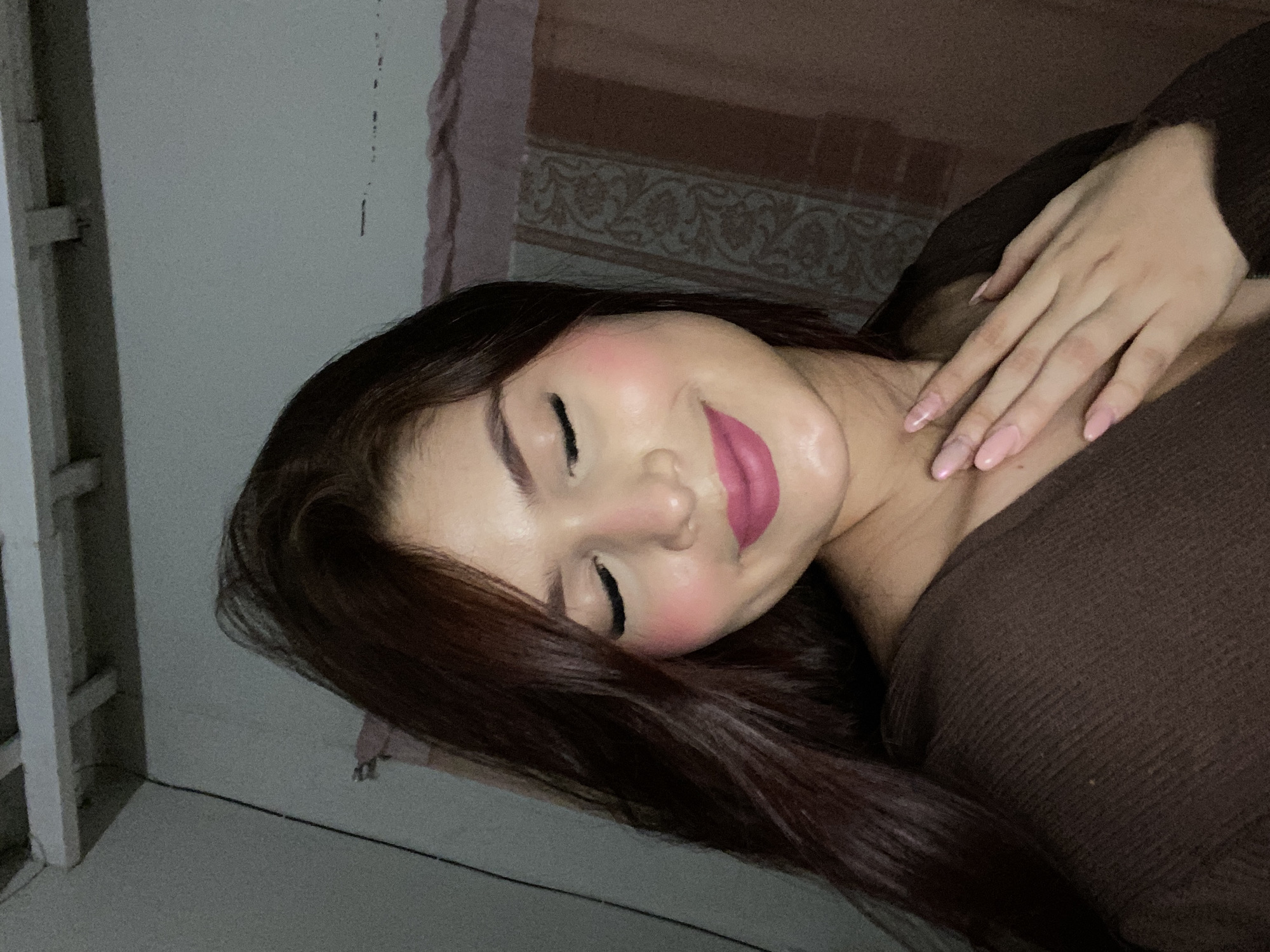 A woman lies on her side with her eyes closed, wearing a brown top. Her right hand rests on her neck. She has long brown hair and pink lipstick. She appears to be indoors, as evidenced by the wall and doorway in the background. The location in Juan Griego, Venezuela, cannot be determined from this image.