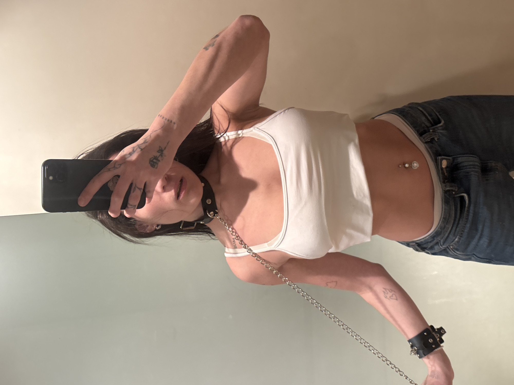 A thin, light-skinned woman with dark hair takes a selfie in a mirror. She is wearing a white tank top and blue jeans. She has a belly button piercing. She has a black leather collar around her neck with a metal chain attached. The chain extends down to her left wrist, which is adorned with a studded black leather cuff. Tattoos are visible on both hands and arms. The background appears to be a wall with light-colored paint. There is no indication of the location being Buenos Aires, Argentina.