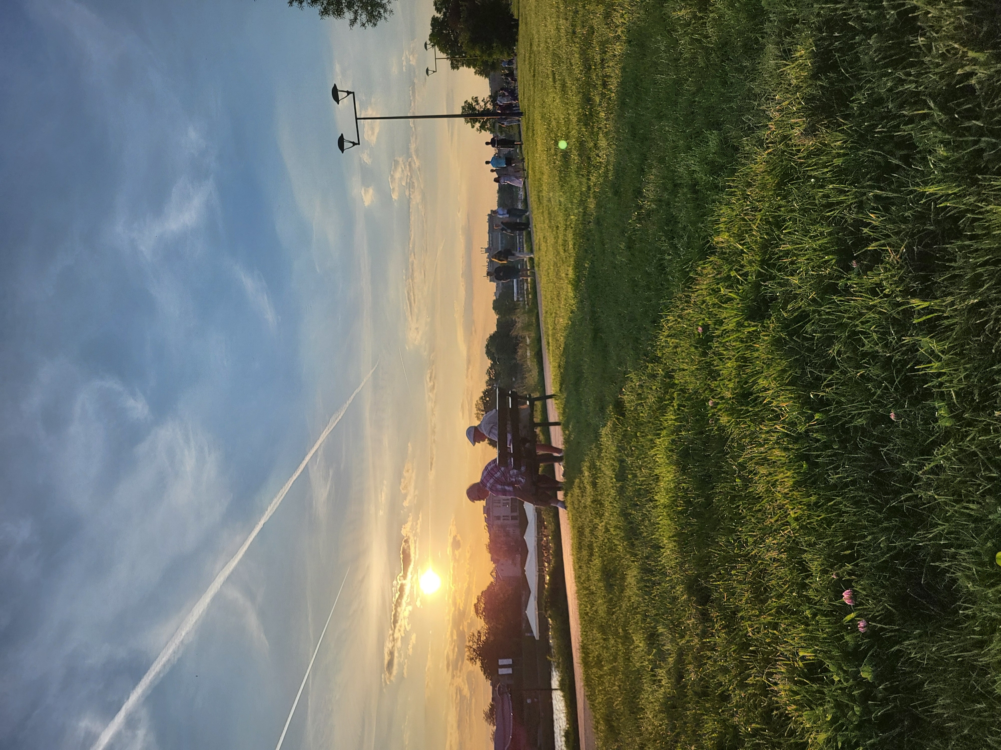 The image depicts a park at sunset. Two people are sitting on a bench. A contrail is visible in the sky. Street lamps are present. Green grass covers the ground. Buildings are in the background.