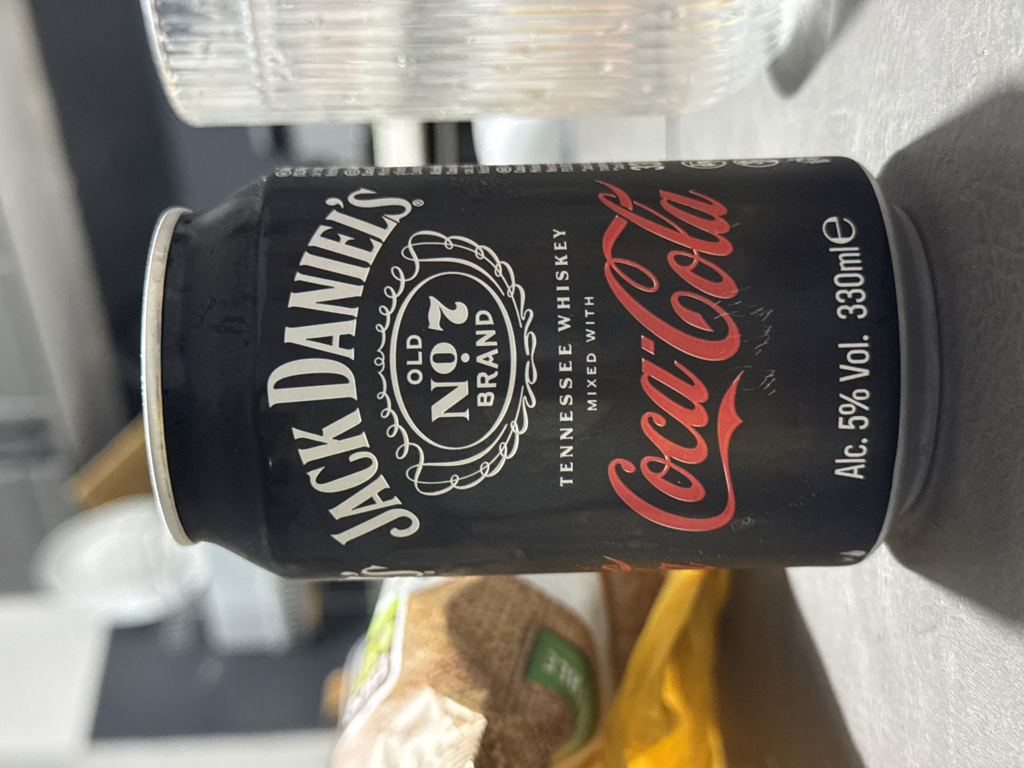 The image displays a close-up shot of a black pre-mixed drink can featuring Jack Daniel's Tennessee Whiskey mixed with Coca-Cola. The can is positioned horizontally on a light grey surface. Prominently visible text on the can includes "JACK DANIEL'S" in white lettering, "OLD NO.7 BRAND" within a detailed oval emblem, and "Coca-Cola" in its iconic red script. Below the primary text, "TENNESSEE WHISKEY" and "MIXED WITH" are stated in white, vertical lettering. On the far right, "Alc. 5% Vol. 330mle" specifies the alcohol content and volume.
In the background, out of focus, a clear, ridged glass or plastic object is visible above the can. Below the can, a yellow item, possibly a bag, and a brown, textured object are partially visible, suggesting other household items or groceries. The overall scene is indoors, likely a kitchen or a similar functional area, with soft, diffused lighting. There is no specific activity depicted; the can is simply resting on the surface. The image was captured in Stara Zagora, Bulgaria. No people are present, and no particular emotions are conveyed.
