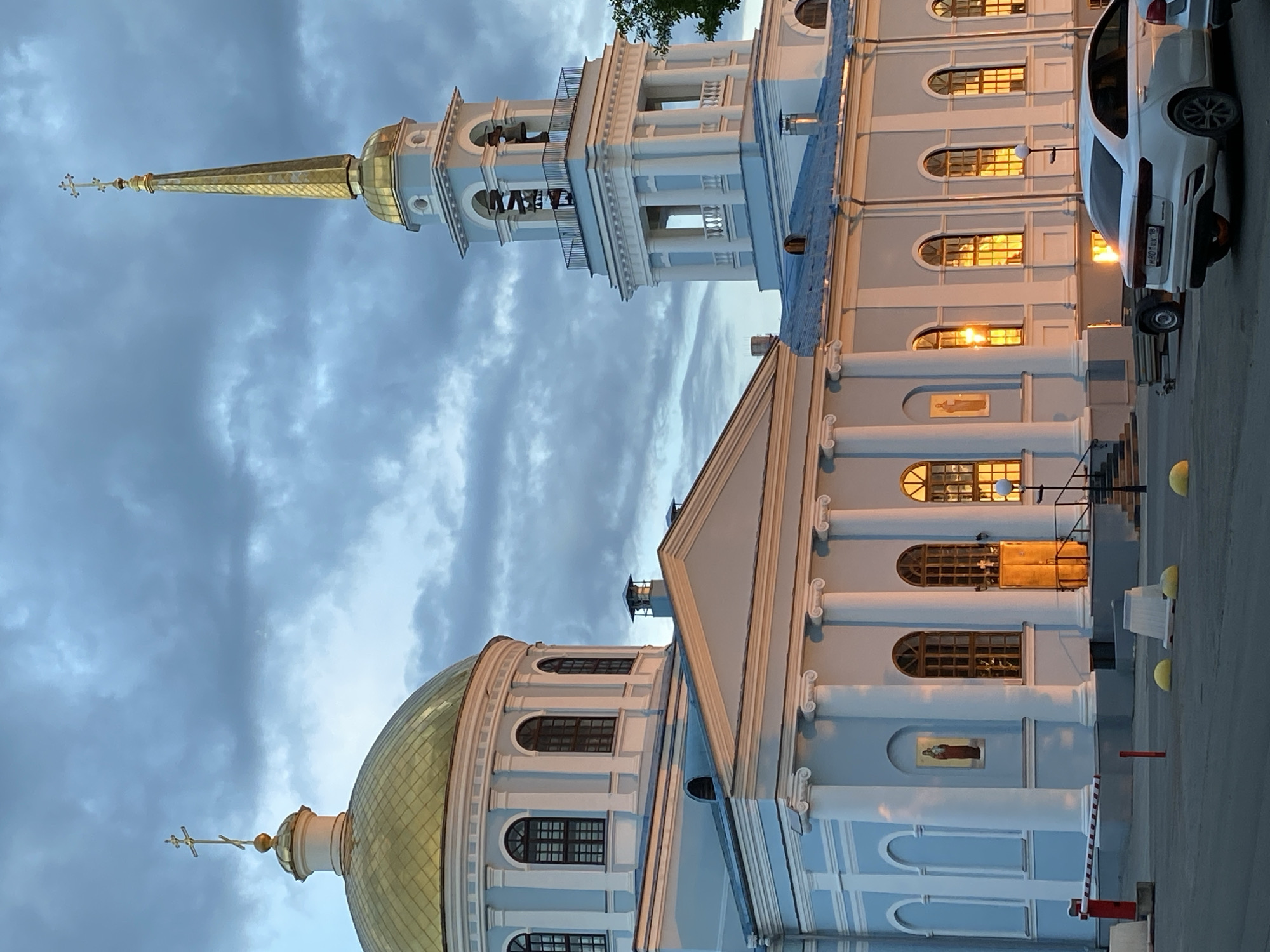 The image depicts the Cathedral of the Annunciation in Votkinsk, Russia. The cathedral has a golden dome on the left and a tall, light-colored bell tower on the top left. The main building has a light-colored façade with arched windows reflecting light. A white car is parked on the right side of the image. The sky is overcast with dark clouds.