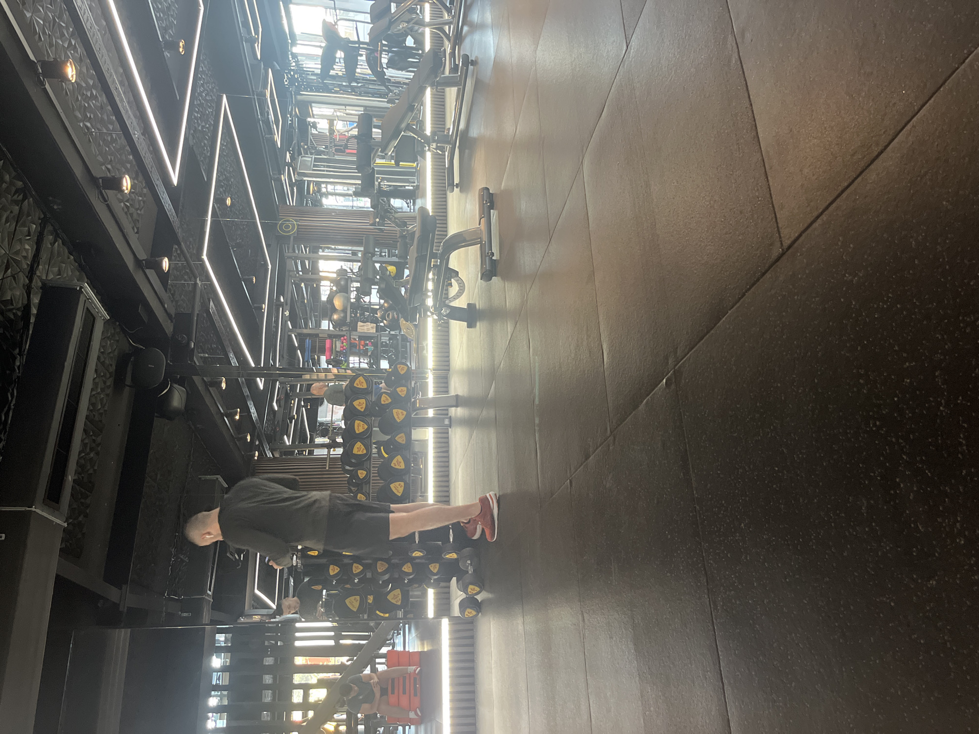 The image shows the interior of a modern gym. There is a man with short gray hair, wearing a long-sleeved gray shirt, black shorts, and red sneakers, exercising in the gym. He appears to be near a dumbbell rack. The gym has a contemporary design with exposed structural elements, metal framework, and integrated lighting. The floor is dark and made of large tiles. Through windows, the outside environment is visible. Given the architecture style and the presence of the equipment, the location is likely an upscale fitness center. It's impossible to confirm the location is in Sofia, Bulgaria without additional context.