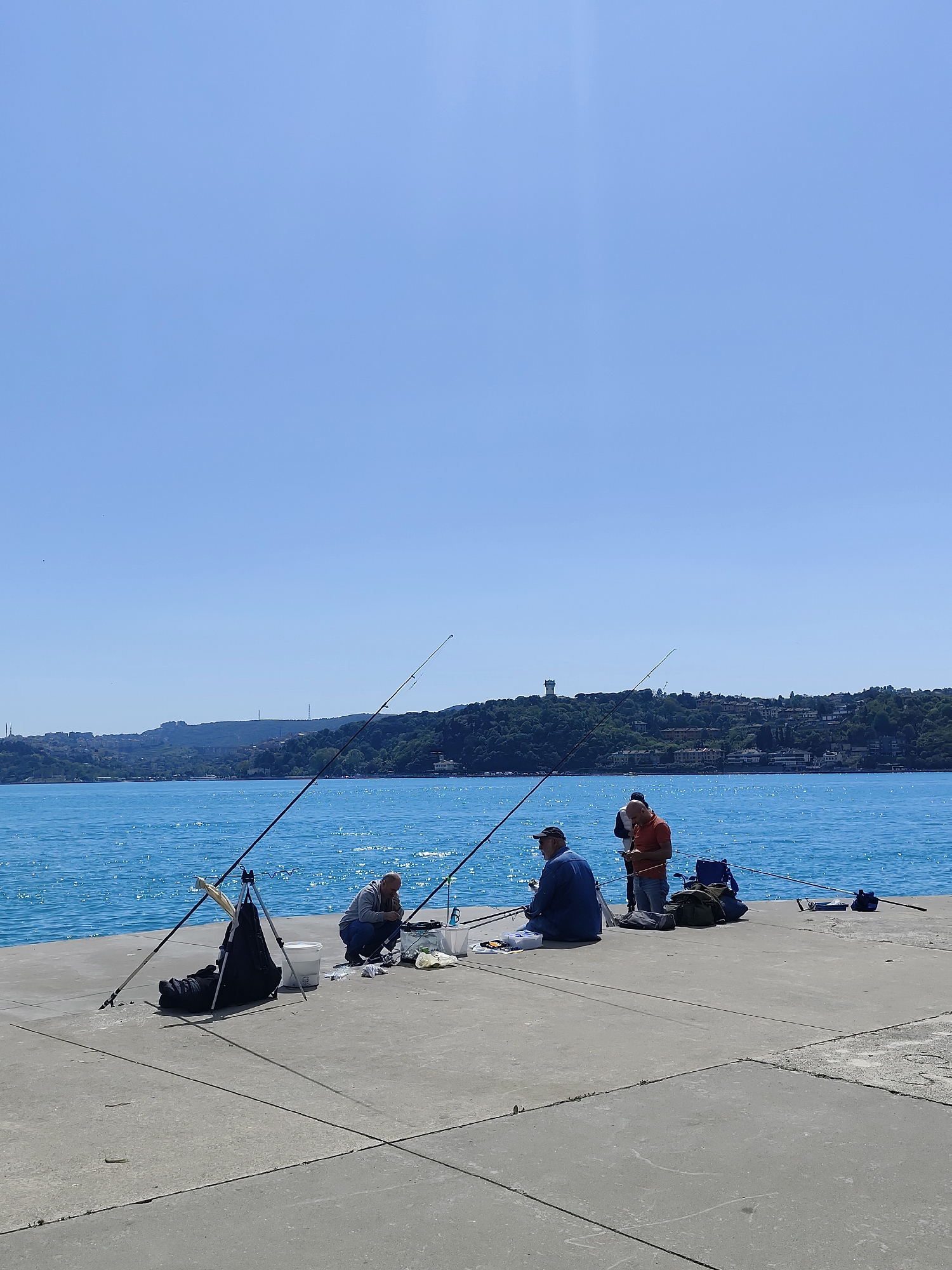 The image shows a group of four people fishing from a concrete pier or dock, on a sunny day. The location is identified as Sarıyer, Turkey, as indicated by the Bosphorus Strait visible in the background, along with the forested hills of the Asian side of Istanbul. The water is a clear blue. Three fishing rods are visible, angling out over the water. The fishermen are positioned along the pier, with fishing equipment scattered nearby. One fisherman is crouched down, seemingly preparing bait or tackle. Another is sitting. A third is standing. The fourth is looking at his cellphone. Items such as fishing bags, buckets, and gear are spread around. The pier surface shows typical concrete block construction.