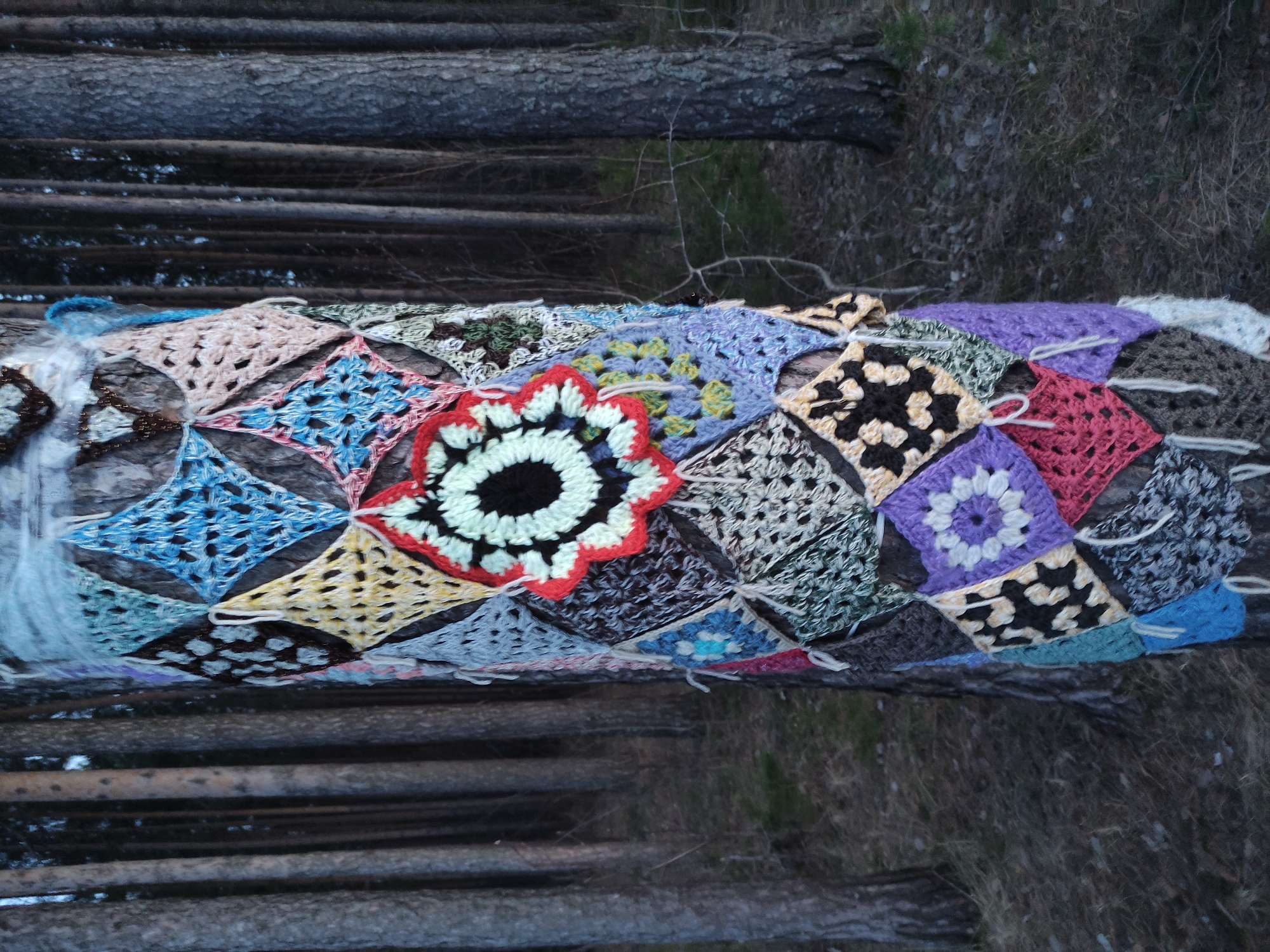 The image shows a colorful crocheted blanket draped over a log in a wooded area. The blanket is made up of various geometric shapes and floral patterns in different colors, including blues, yellows, purples, reds, and blacks. The log is part of a fallen tree trunk, and the background consists of other trees and vegetation. The setting appears to be a forest or park-like area. There is no indication that this is in Chaikovsky, Russia.
