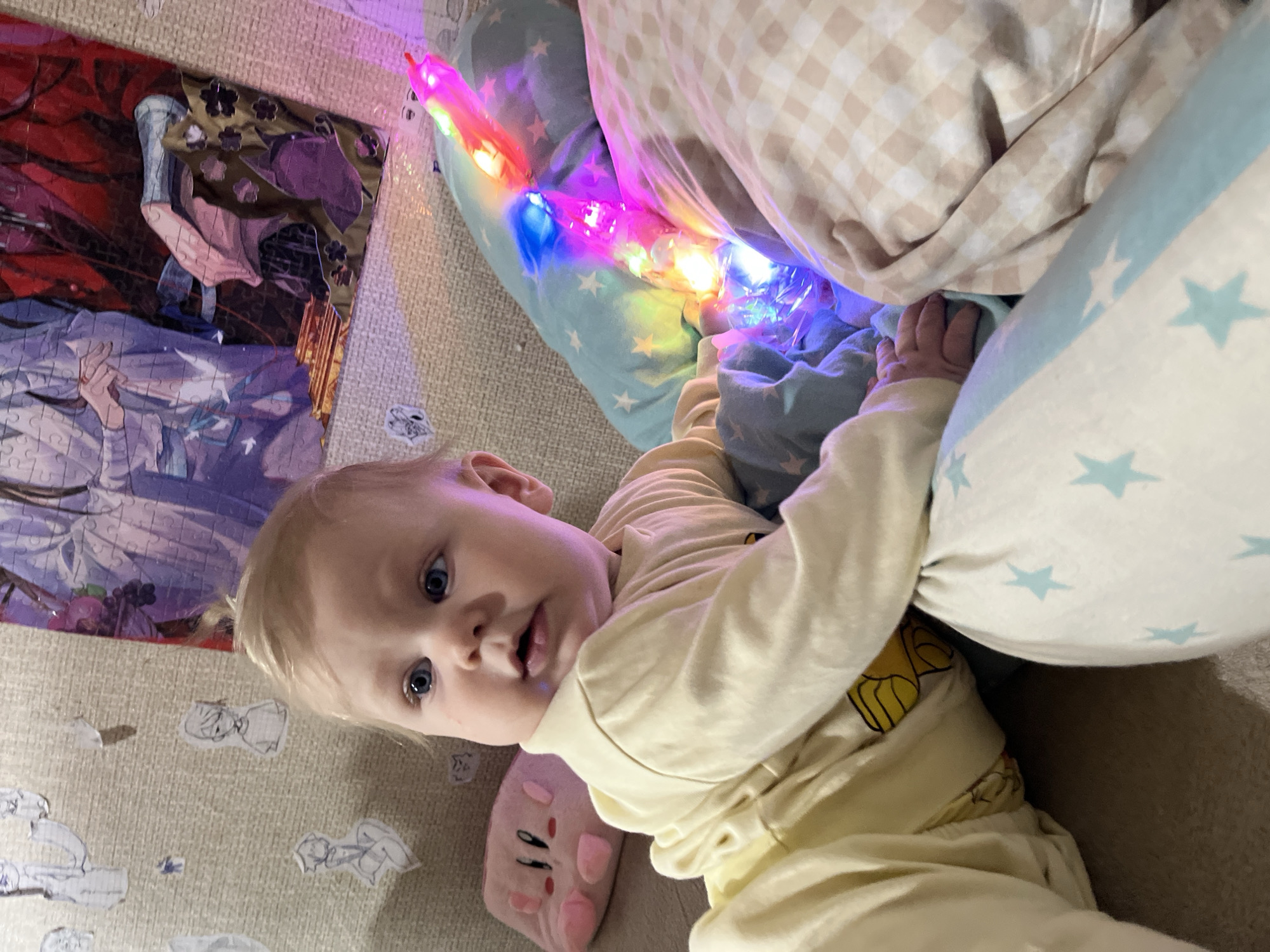An indoor, portrait-oriented shot captures a baby lying down and looking directly at the camera in Vesyoloye, Russia. The baby, with fair skin, light blonde hair, and wide blue eyes, wears a light yellow long-sleeved top with a yellow cartoon character design featuring ears and rosy cheeks. The baby's expression is one of calm curiosity.
To the baby's right, a light blue pillow decorated with white stars is positioned, alongside a beige and white checkered blanket. Nestled between the baby and the pillow is a vibrant, multi-colored light-up toy or decoration, possibly a wand, emitting red, blue, green, and pink light.
Behind the baby's head and to the left, a partially completed jigsaw puzzle is visible on a light-colored carpet or rug. The puzzle depicts anime-style characters, including one with flowing purple and white garments and another figure wearing a dark purple hat. Several small, hand-drawn paper cutouts are scattered on the carpet near the puzzle. A pink plush toy, resembling Kirby, is partially visible near the baby's left arm. The scene conveys a cozy and playful domestic atmosphere, likely in a living room or bedroom. No specific time of day or outdoor weather can be determined, and no discernible text is present in the image.