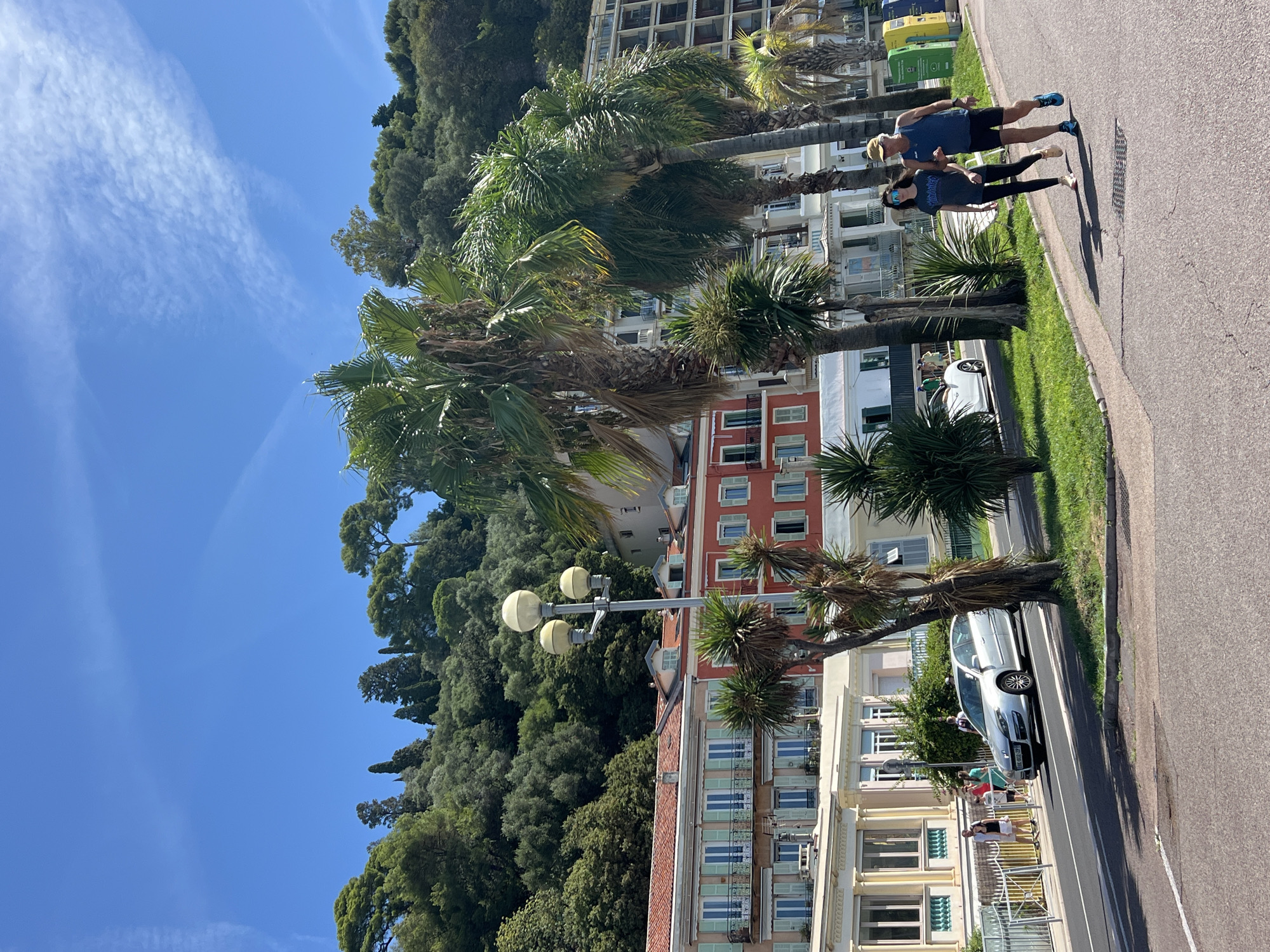 The image depicts a sunny day on the Promenade des Anglais in Nice, France. In the foreground, a man and a young woman are walking on a paved path. The man wears a blue tank top, dark shorts, and a yellow cap, while the woman has on a grey t-shirt, black leggings, and blue sunglasses. Behind them, several palm trees line the path. Further back, along the waterfront, a silver car is parked on the left side of the road. Several buildings with balconies and shutters are visible, painted in shades of cream, coral, and red. In the background, a steep, tree-covered hillside rises behind the buildings, under a clear blue sky with wispy clouds. The overall scene suggests a leisurely stroll in a picturesque coastal city.