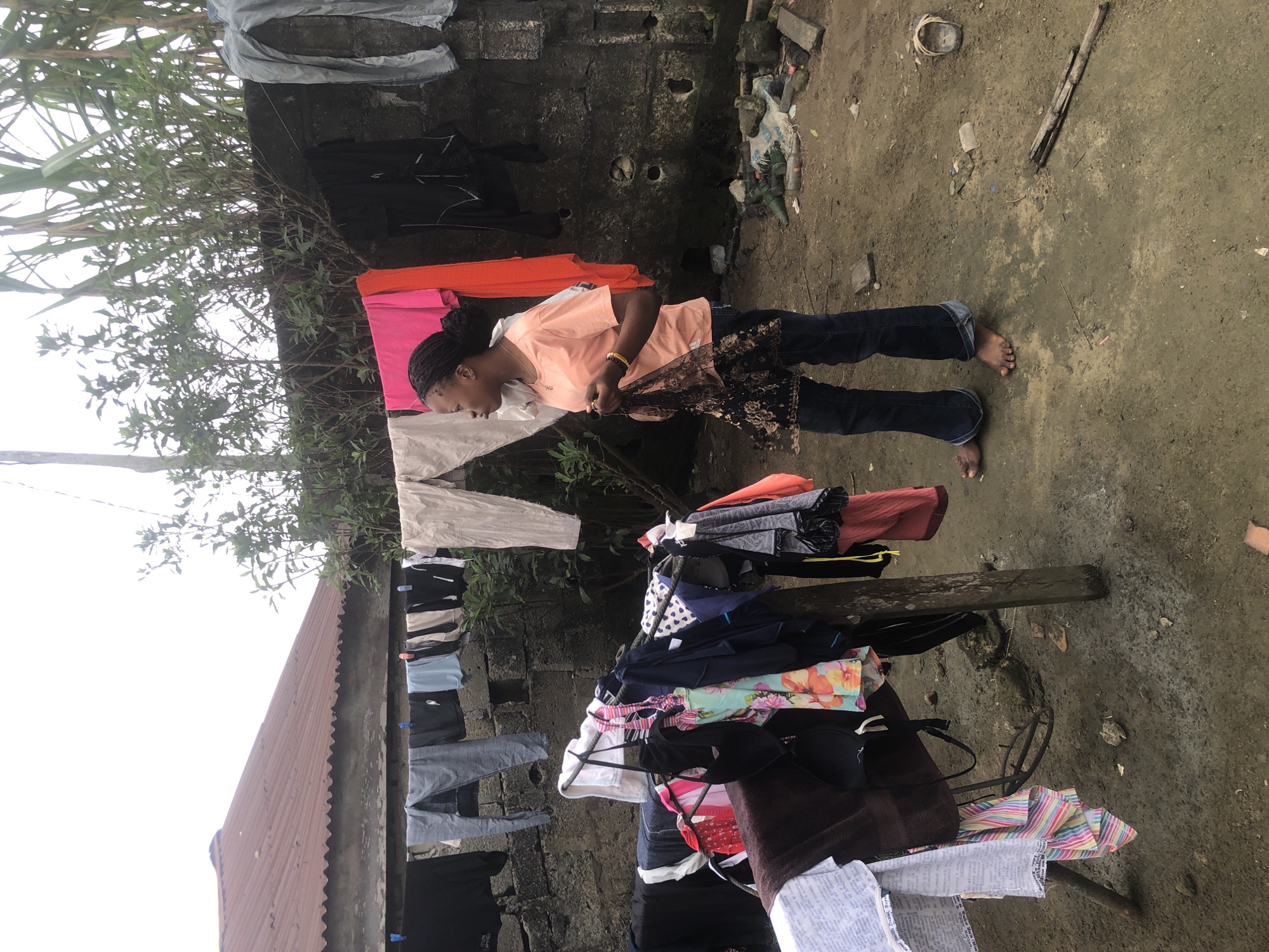 A young Black woman stands barefoot on a dirt ground in Ado, Nigeria, near a clothesline with laundry hanging. She is wearing a peach-colored t-shirt and dark blue jeans. She holds a black, patterned fabric in her hands. The background includes a concrete wall, a corrugated metal roof, and overgrown vegetation. The ground is uneven and appears damp, with scattered debris including a worn sandal, sticks, and what appears to be some discarded items. The scene suggests a residential outdoor space where laundry is being done.