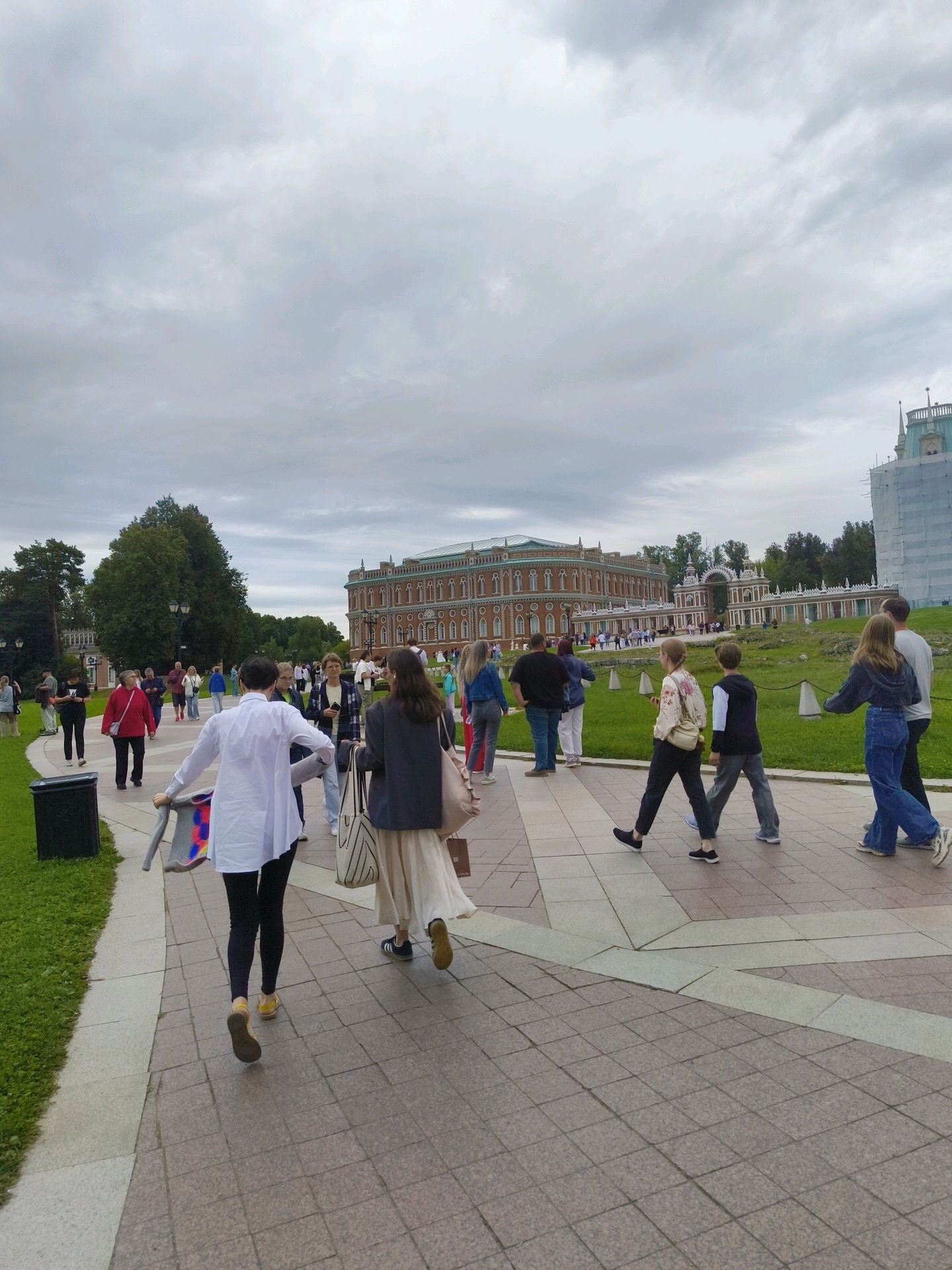 A medium-shot of a group of people walking on a paved path in what appears to be a park with a large ornate building in the background. The building is made of brick with white trim and has a distinctive architectural style. The sky is overcast. A few trees are visible on the left side of the image, and a fence separates the path from a grassy area. The people are dressed in casual clothing, and some are carrying bags. The paved path is made of square tiles with light-colored lines forming a pattern. The image is taken from a slightly elevated perspective, looking down at the path. The location is Tsaritsyno Park in Moscow, Russia, identifiable by the architecture of the Grand Palace and the park's layout.