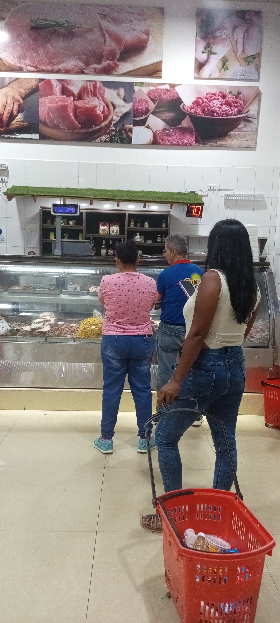 The scene captures three individuals at a meat counter in a grocery store. Two women and one man are present. The woman closest to the camera is pulling a red shopping basket containing assorted items. The other woman, wearing a pink shirt with star patterns, and the man, in a blue shirt, are directly facing the meat display counter. Above the counter, a display shows various cuts of meat, and a digital display reads "70." Shelves above the counter contain various bottles and jars. The environment appears to be a typical grocery store setting, with a tiled floor and white-tiled walls.