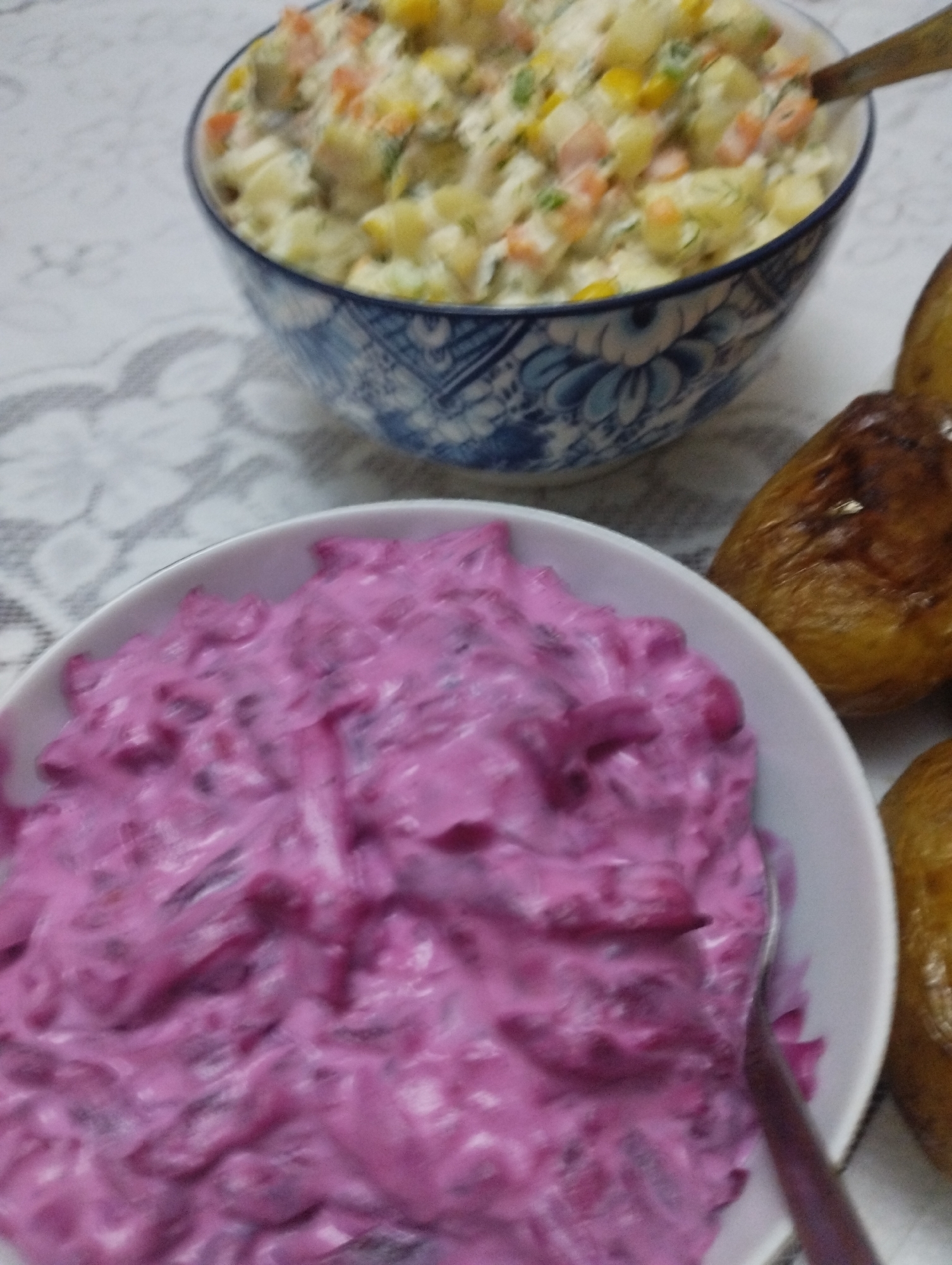 The image shows two bowls of food and several baked potatoes on a tablecloth. The bowl in the foreground contains a vibrant pink salad, possibly made with beets and a creamy dressing, with a spoon partially submerged in it. To the upper right of this bowl, another bowl contains a lighter-colored salad with visible pieces of vegetables like corn and carrots, also with a spoon resting on the edge. To the right of the bowls, several whole baked potatoes with browned skins are arranged. The scene is set indoors, likely on a table covered with a patterned tablecloth. There are no people visible in the image. The lighting appears to be natural, coming from above and slightly to the right. No specific location within Bilajari, Azerbaijan, can be identified from the visual information provided.