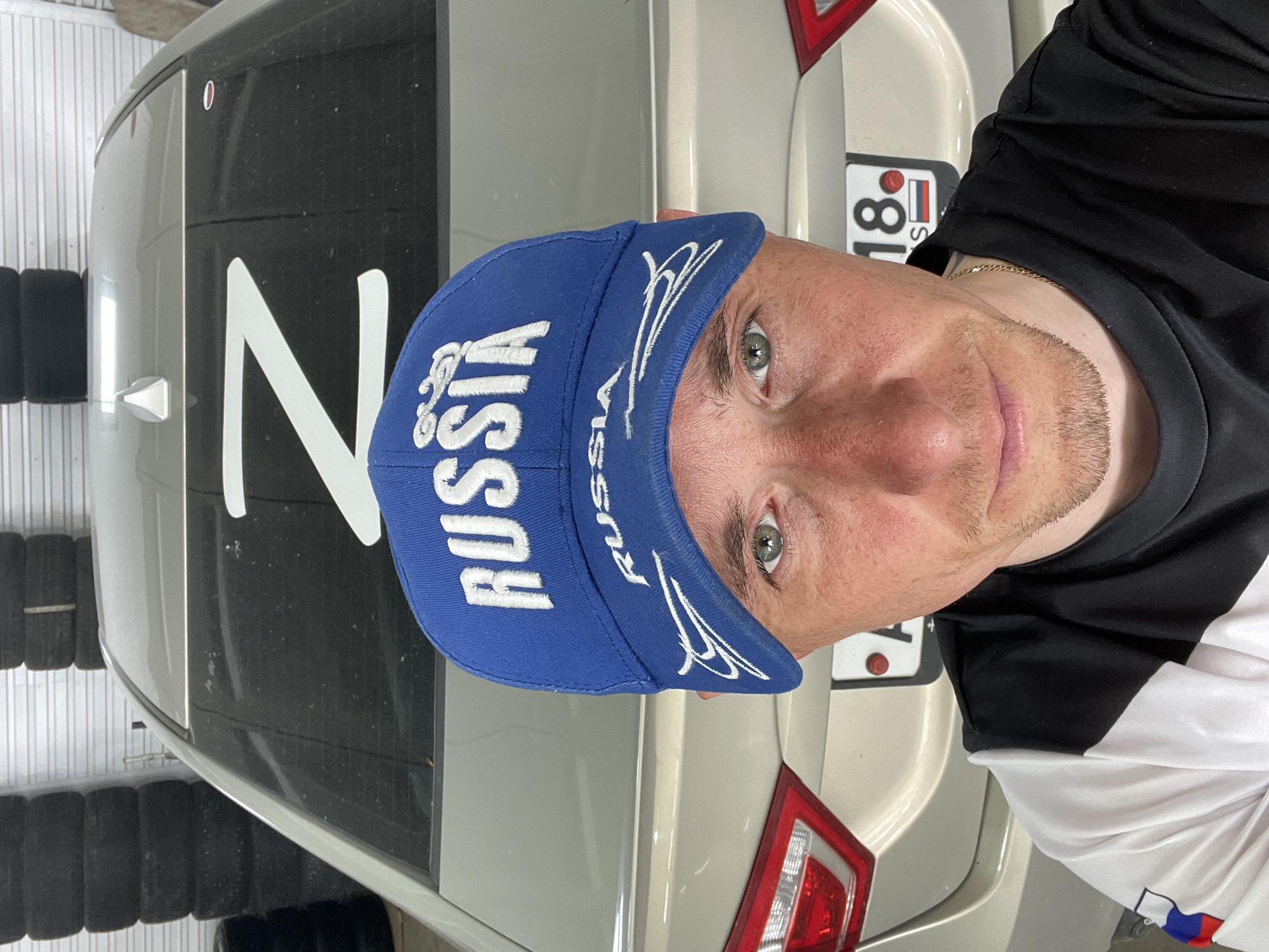 A close-up selfie shot features a man wearing a blue baseball cap with "RUSSIA" embroidered on it, positioned in front of a light-colored vehicle, possibly a car. The rear of the vehicle is visible, displaying a large white "Z" symbol and a license plate partially visible with the characters "18" and the Russian flag. Tires are stacked behind the vehicle on the left side. The man is wearing a black and white t-shirt with a gold chain around his neck. The location is identified by the license plate as being in Sarapul, Russia.