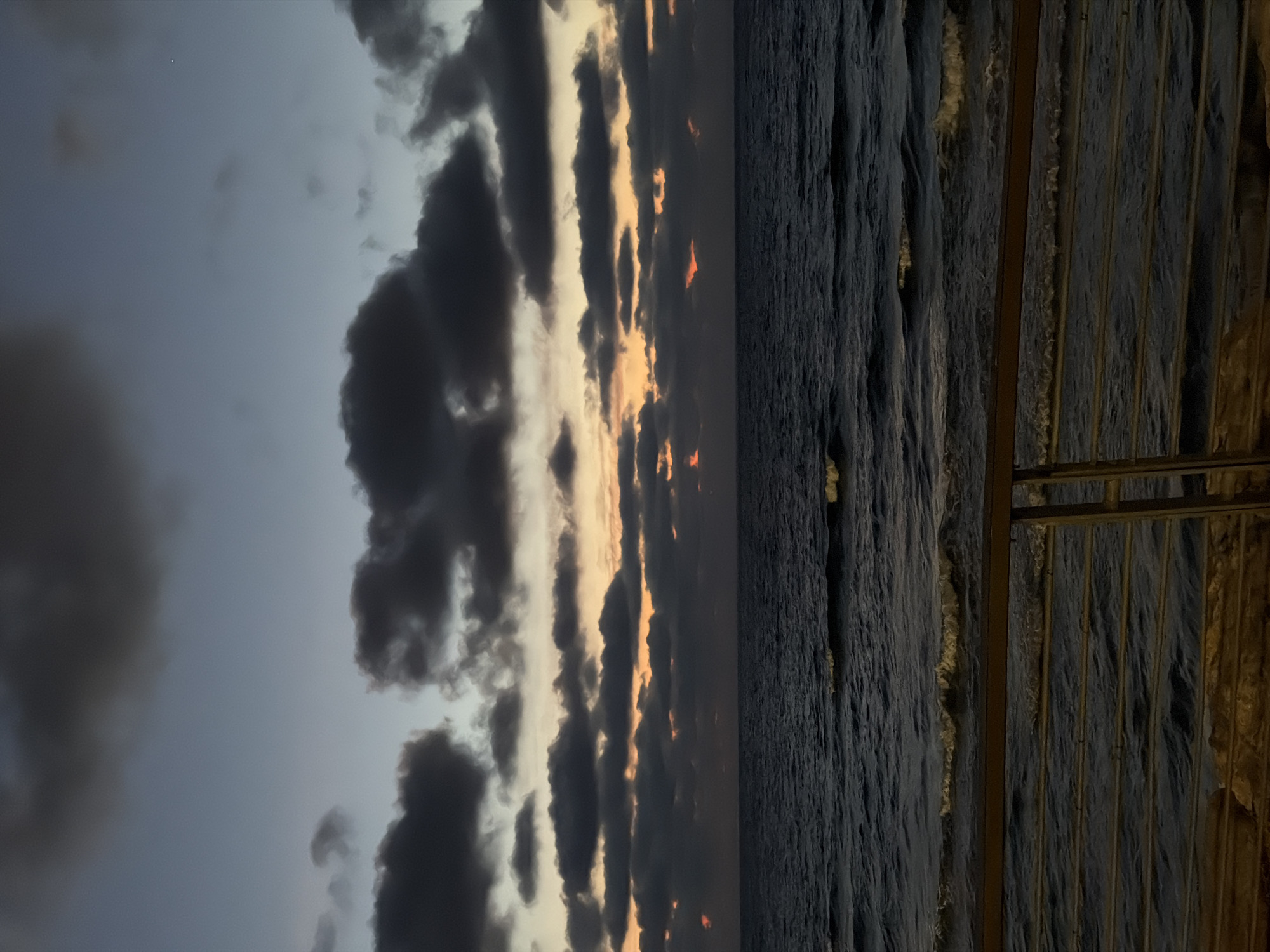 The image captures a view of the sea in Tel Aviv, Israel, at dusk or dawn. The sky is filled with dark, ominous clouds illuminated by the faint light of the rising or setting sun. The water is dark and choppy with evident waves. A metal railing runs along the right side of the frame, suggesting the vantage point is a balcony or elevated walkway, partially obscuring the view of the water. There are no people visible.
