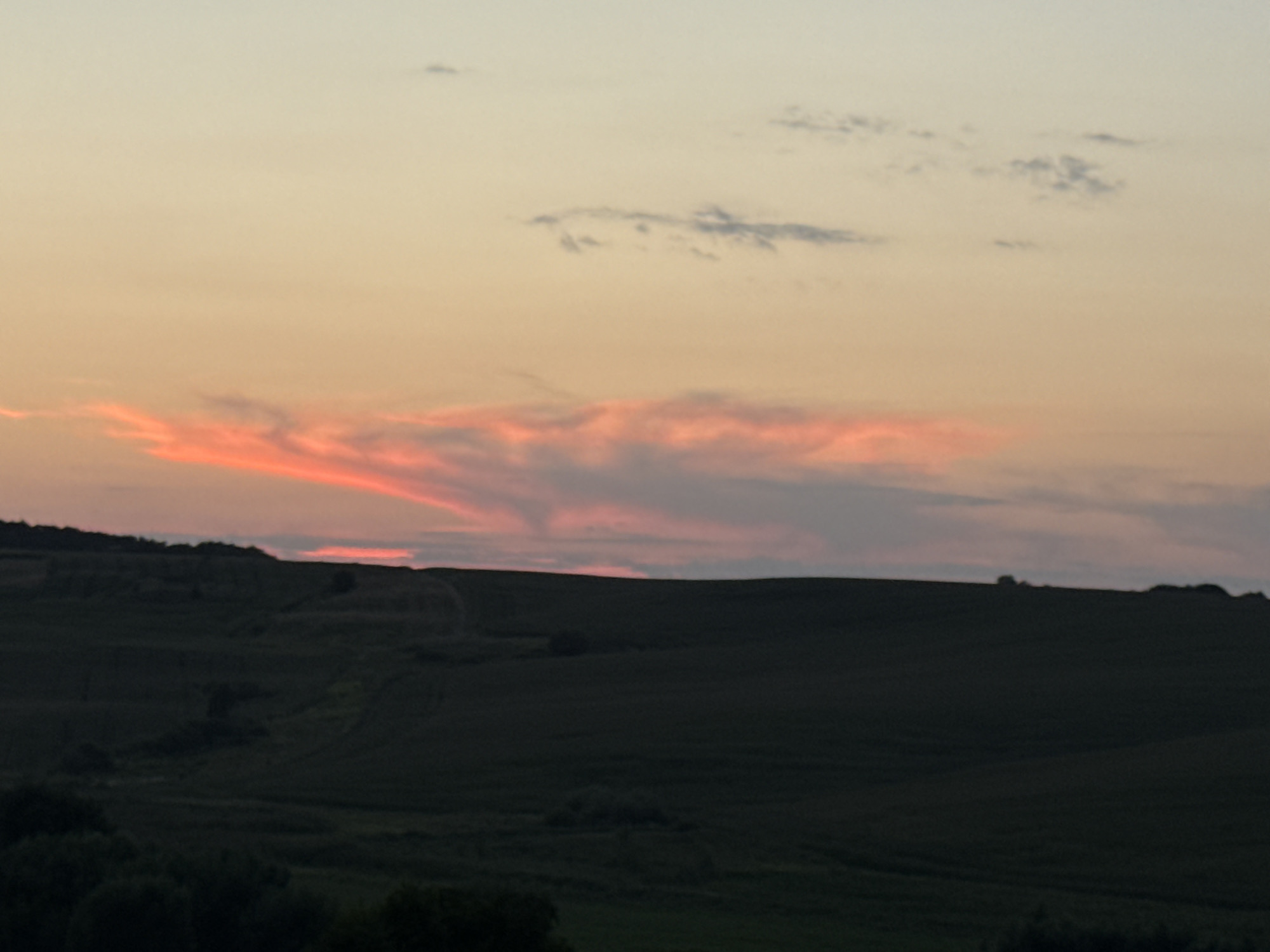 A landscape photograph captures rolling, dark green hills under a sky painted with the warm hues of sunset. The sky transitions from a pale yellow at the top to a soft orange and pink near the horizon, where wispy clouds are tinged with vibrant pink. The hills are densely covered in vegetation, appearing as a dark silhouette against the lighter sky. Some scattered trees or bushes are visible on the hillsides. The overall mood is serene and tranquil, indicative of dusk in a rural or semi-rural setting.