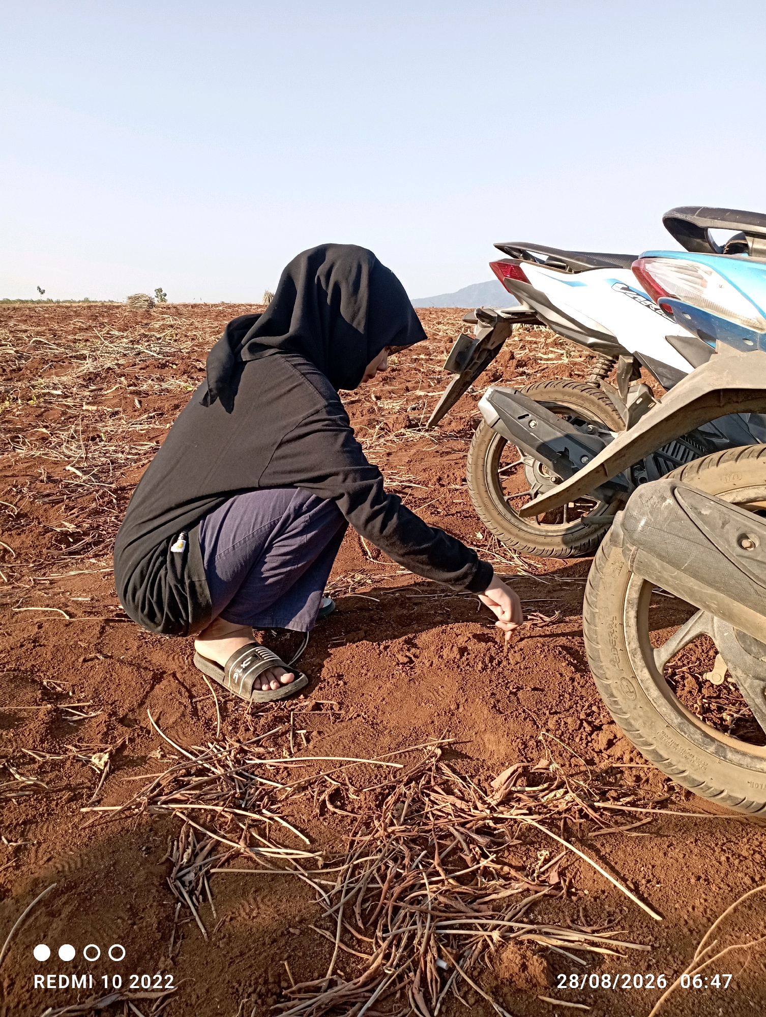 A person wearing a black hijab and a black hoodie is crouching on dry, reddish-brown soil. The person is reaching down with their right hand to touch something on the ground. The environment appears to be an open field with sparse dry vegetation. Two motorcycles are parked nearby, one partially visible in the foreground and another in the background. The sky is clear and blue. The timestamp "REDMI 10 2022" is visible in the bottom left corner, and the date and time "28/08/2026 06:47" are in the bottom right corner. The location is identified as Gandik, Indonesia.