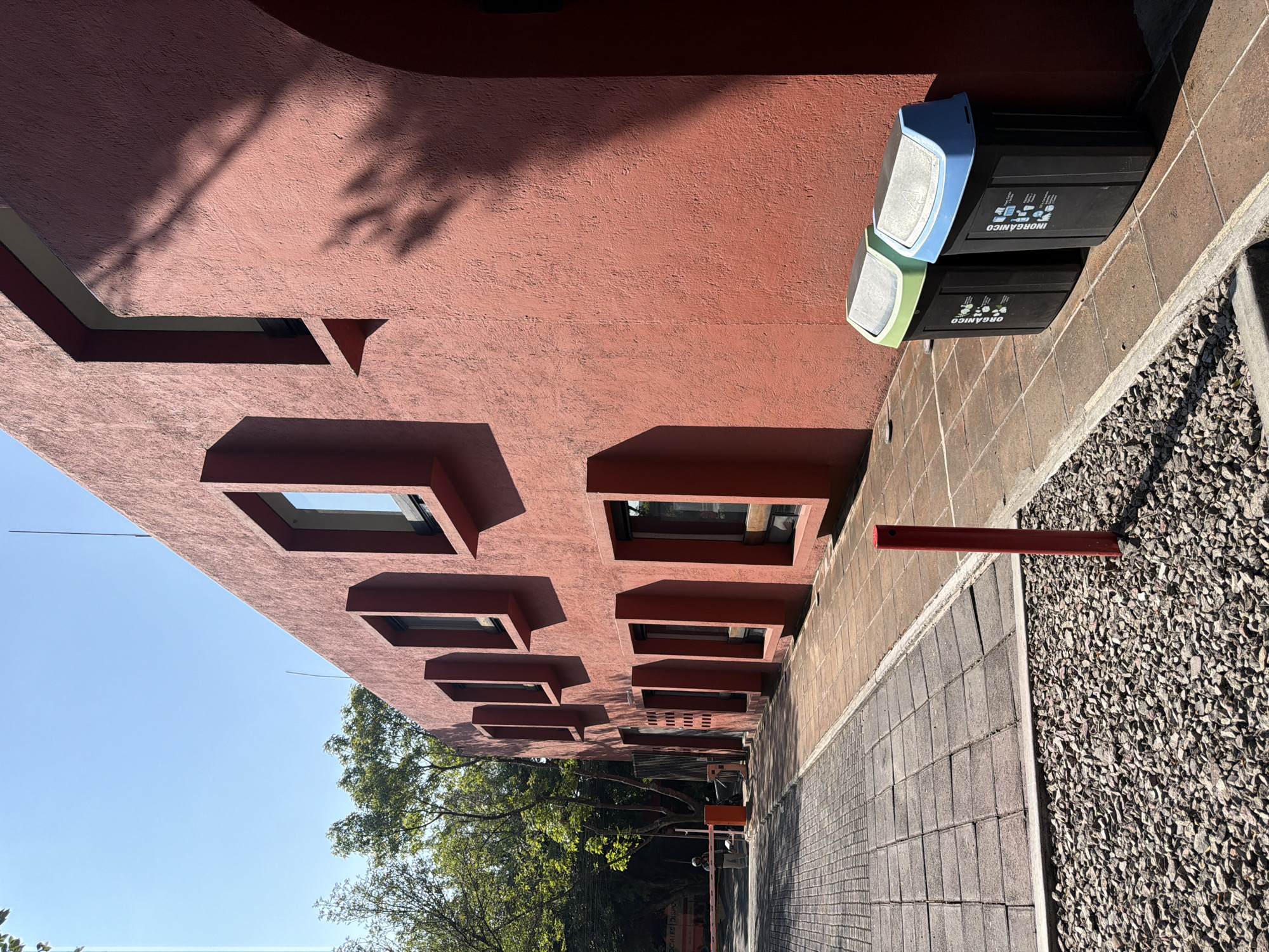 The image shows a section of a building in Puebla, Mexico. The building's exterior is a deep reddish-brown color with a textured surface. There is a row of square-shaped, recessed windows along the side of the building, which are rectangular frames with windows within. The bottom edge of the image shows trees and a clear blue sky. To the right of the building is a sidewalk made of light gray blocks. A section of gravel extends from the sidewalk towards the edge of the image. At the top right of the image, there are two trash bins with blue and green accents. The trash bins have markings indicating the type of waste they are intended for ("INORGÁNICO" and "ORGÁNICO"). A red metal pipe extends from the building out into the gravel portion.