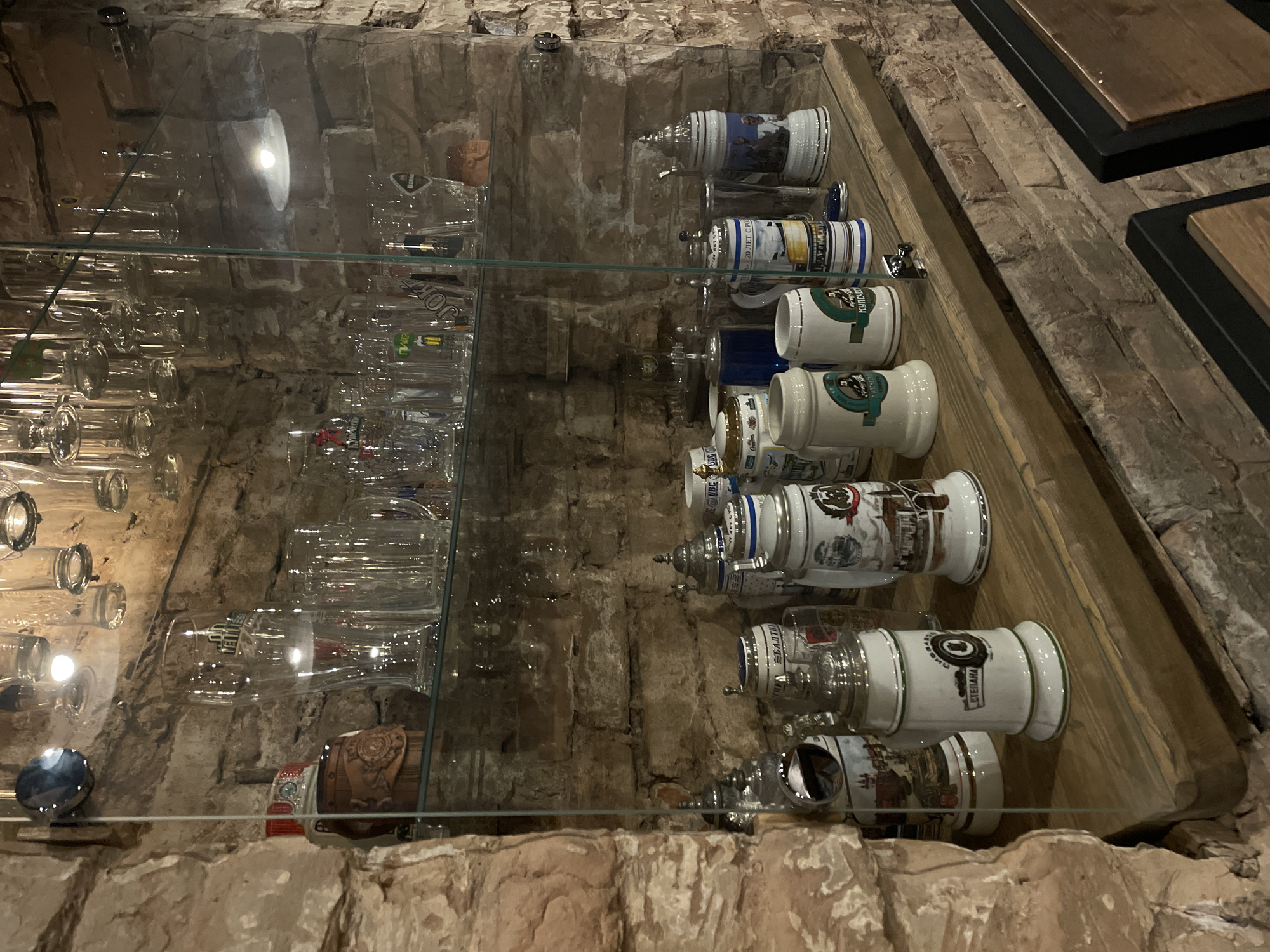The image shows a display case in Krasnoyarsk, Russia. The case contains rows of clear glass drinking glasses on the left and decorative ceramic beer steins on the right, all organized on wooden shelves. The background is a brick wall.