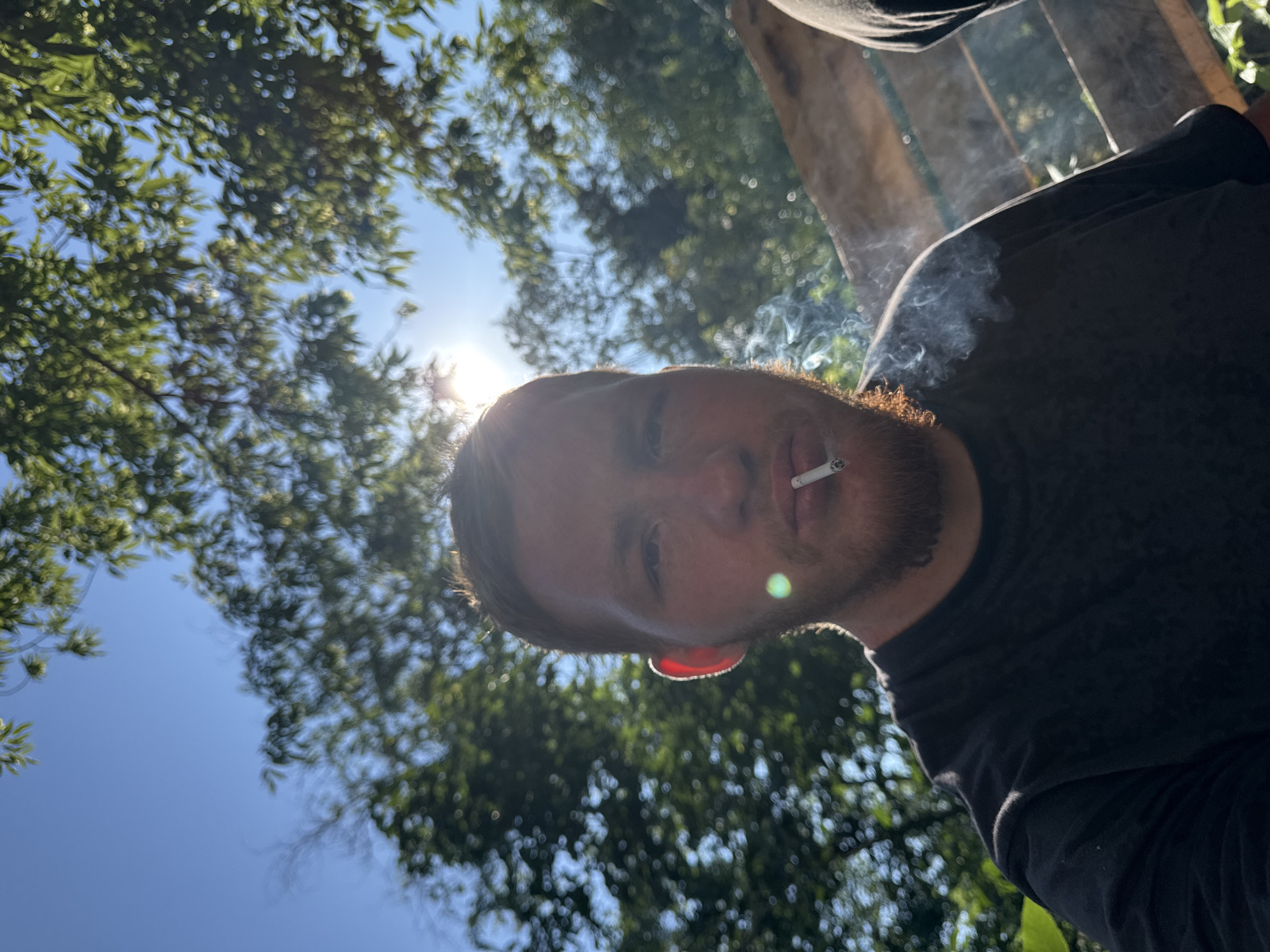 An outdoor, eye-level, medium shot shows a Caucasian man with red hair and a beard, wearing a black t-shirt, smoking a cigarette. The man is positioned in the center of the frame, looking directly at the camera. Sunlight filters through green foliage, creating a dappled effect and lens flare. The background consists of out-of-focus trees and a wooden structure. The man's face is partially illuminated by the sun, with the light creating a bright rim around his ear. Smoke rises from the cigarette and dissipates around his face. Another person, out of focus, is visible to the right of the frame.