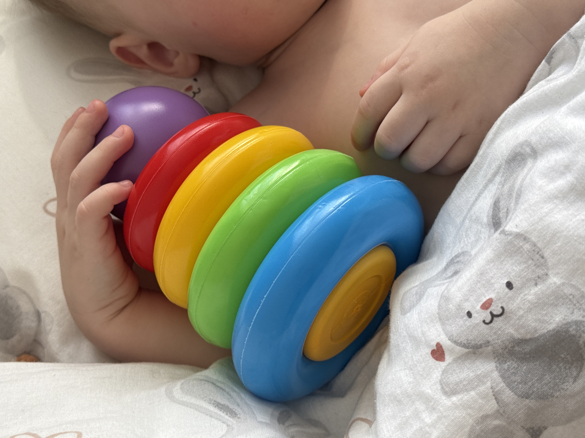 An infant is lying on a white blanket with a gray bunny pattern. The infant is holding a multi-colored stacking toy consisting of purple, red, yellow, green, and blue rings. The location is unknown and cannot be determined from the image.