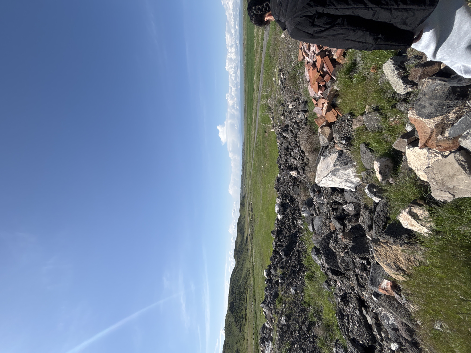 The image presents a scene from Suser, Armenia, showcasing a natural landscape juxtaposed with human presence. In the foreground, a rough terrain of rocks, grass, and scattered red bricks dominates the view. A person wearing a black jacket and light-colored pants partially occupies the upper right corner of the frame, implying an observer's viewpoint. Beyond the rocky foreground, a vast expanse of green fields stretches towards the horizon. The clear blue sky occupies a significant portion of the left side of the image, dotted with a few white clouds near the horizon. The overall environment suggests an elevated vantage point overlooking a rural area in Suser.